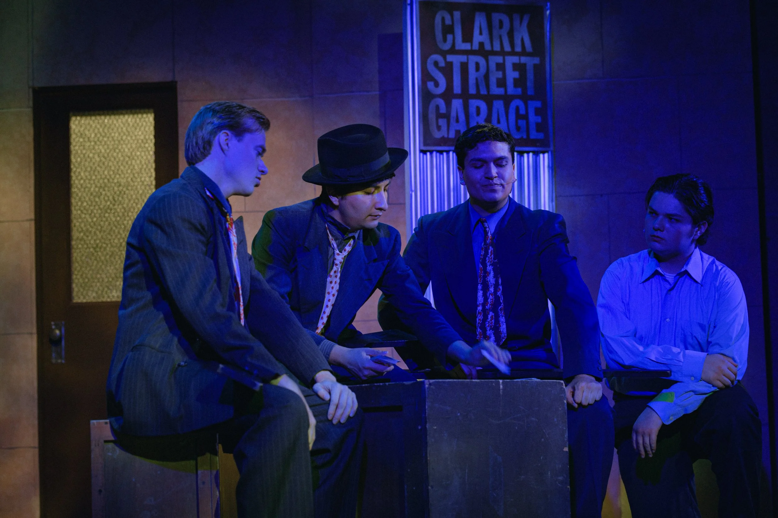 Four actors in a stage performance, wearing suits and ties, with a "Clark Street Garage" sign in the background. One actor wears a hat and they appear engaged in dialogue or a scene, set in a dimly lit, theatrical environment with blue lighting.