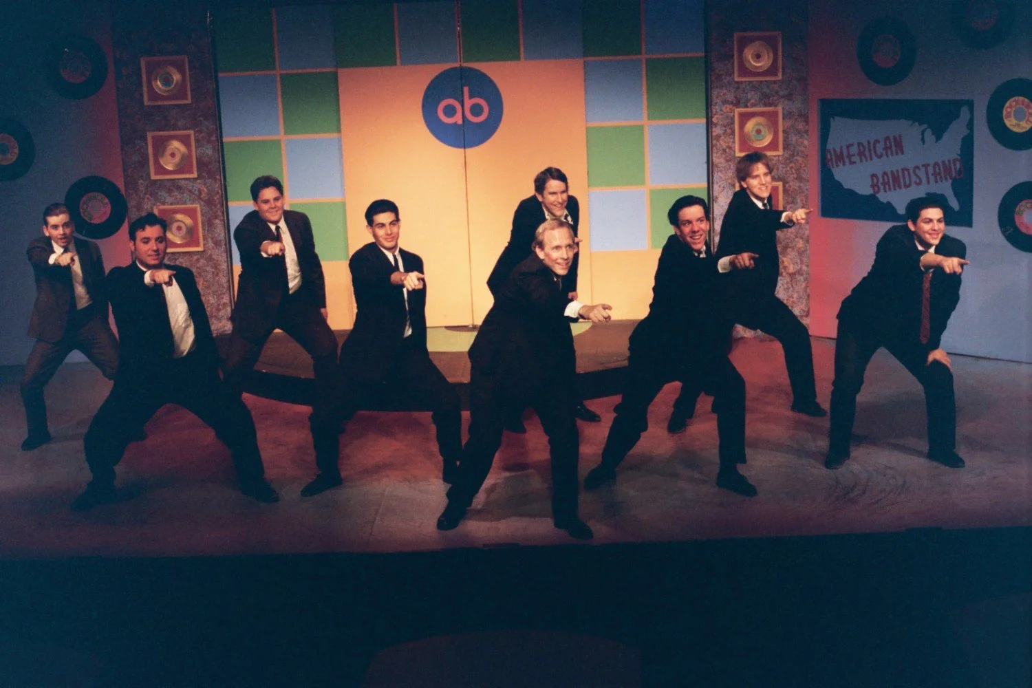 A group of nine men dressed in suits performing a choreographed dance on stage in front of an "American Bandstand" backdrop with colorful decor and vinyl records on the walls.