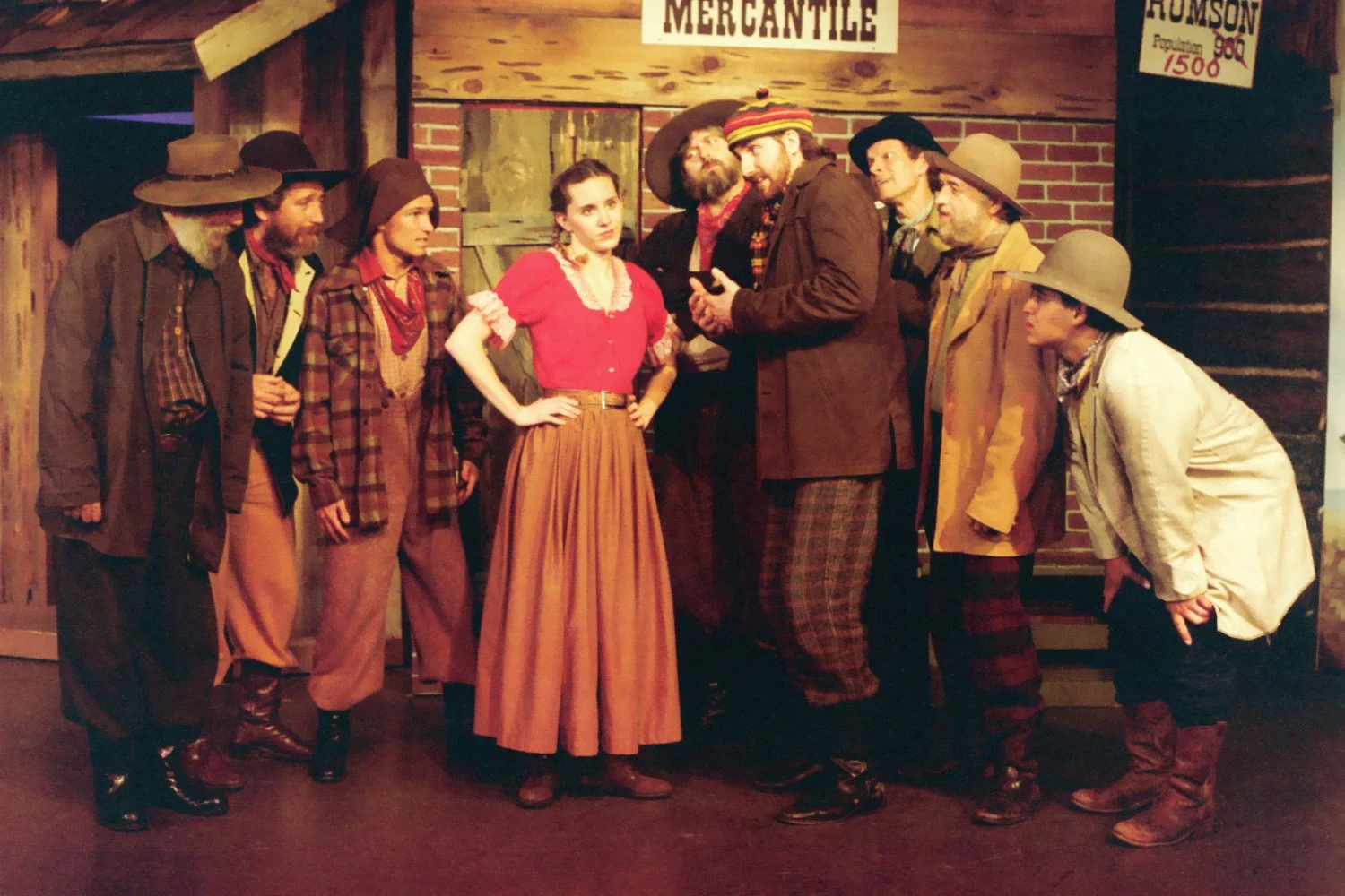 Group of people dressed in Western-style clothing gathered on a stage set resembling a rustic town scene.