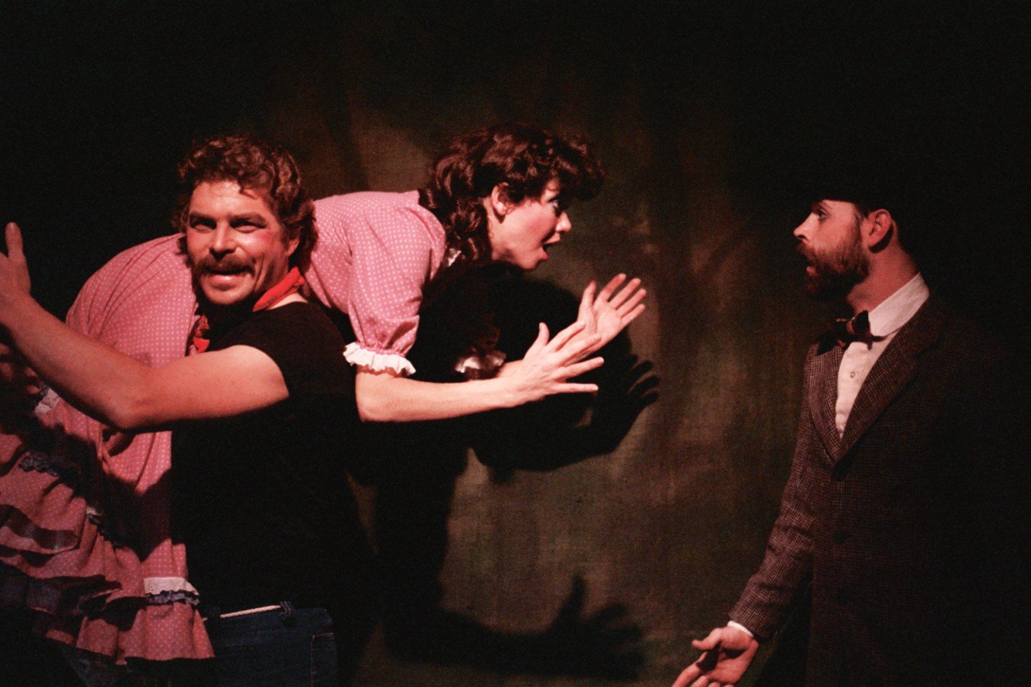 A man with a mustache playfully carries a woman in a pink dress on his shoulder, while another man in a suit looks on with surprise.