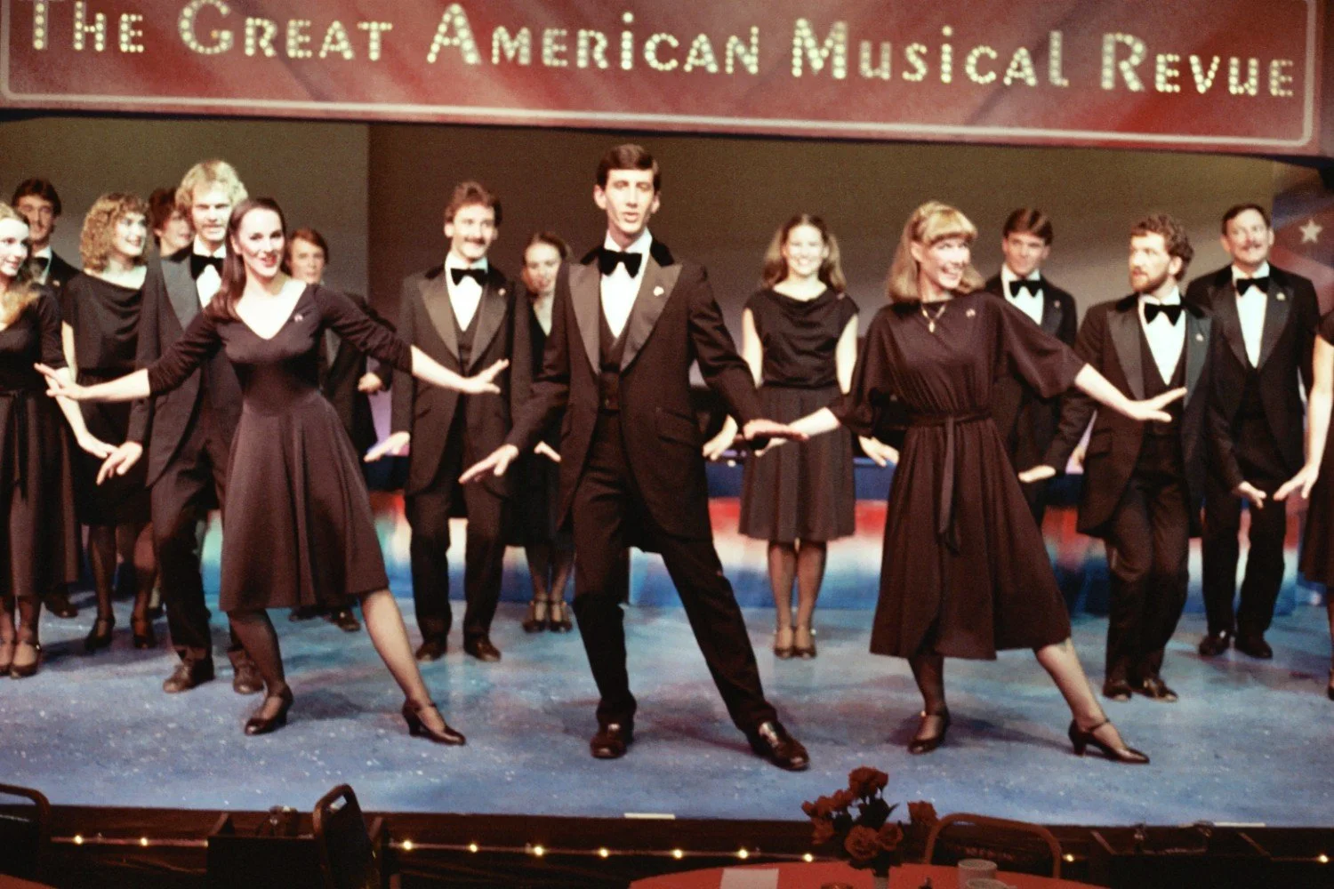 Group of performers in formal attire dancing on stage under a sign that reads "The Great American Musical Revue."