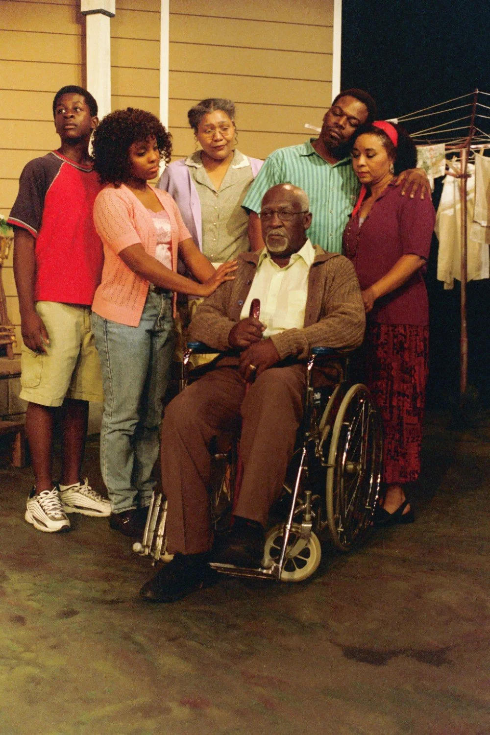 A group of people surrounding an elderly man in a wheelchair in a domestic setting.