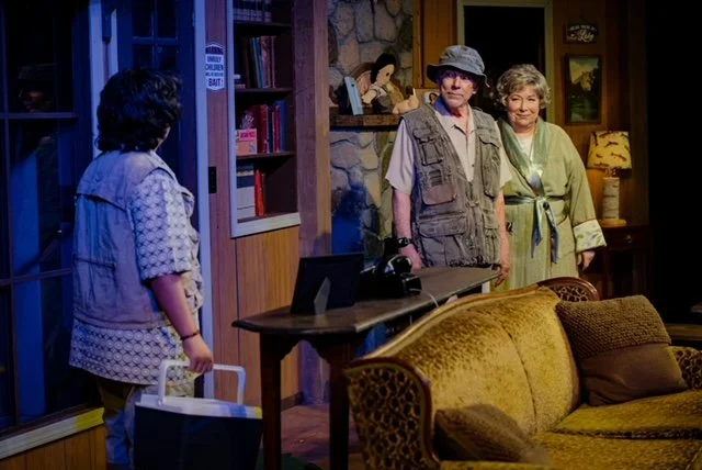 Three people in a living room setting, two standing in the background near a doorway and one facing them holding a bag. The room has wooden walls, a sofa, bookshelves, and decorative items. Warm lighting creates a cozy atmosphere.