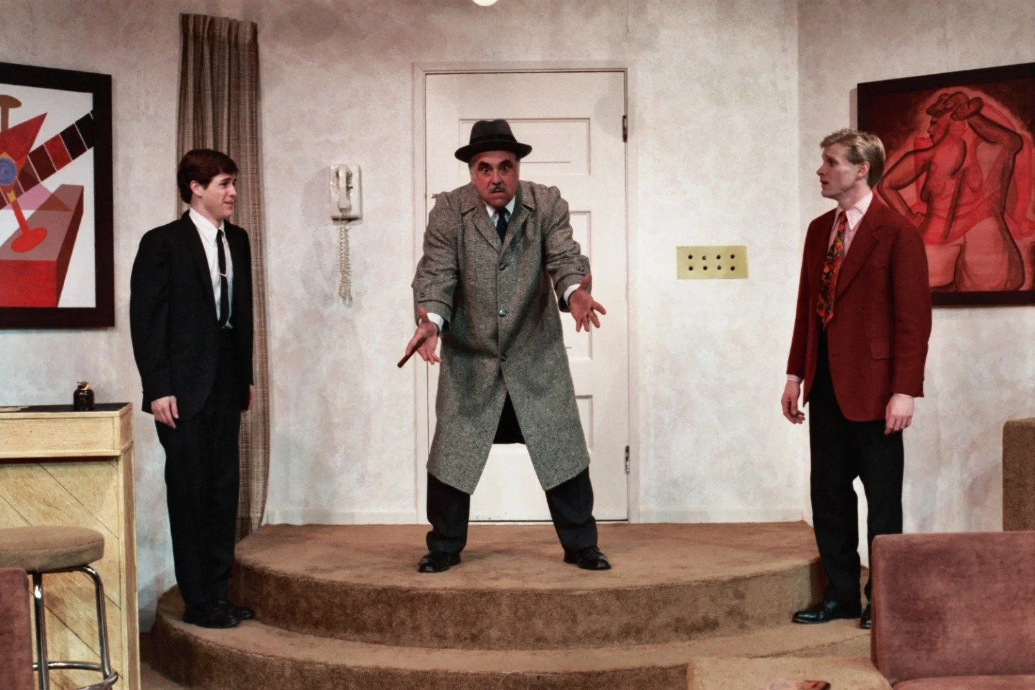 Three men in a living room setting, one gesturing dramatically. The men are wearing suits and standing on steps. Abstract paintings are visible on the walls.