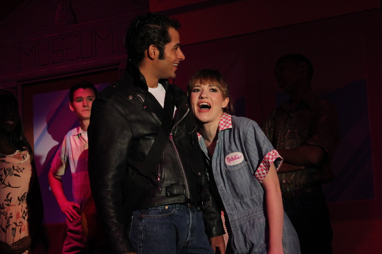 Stage performance with a man in a leather jacket and a woman in a mechanic jumpsuit labeled "Natalie." Others stand in the background against a museum backdrop.