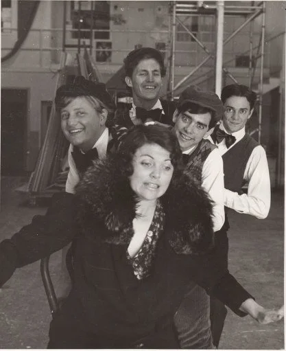 Vintage black and white photo of five people in playful poses on a set, with one woman in front wearing a fur stole and four men behind her dressed in vintage-style clothing, possibly theatrical performers.