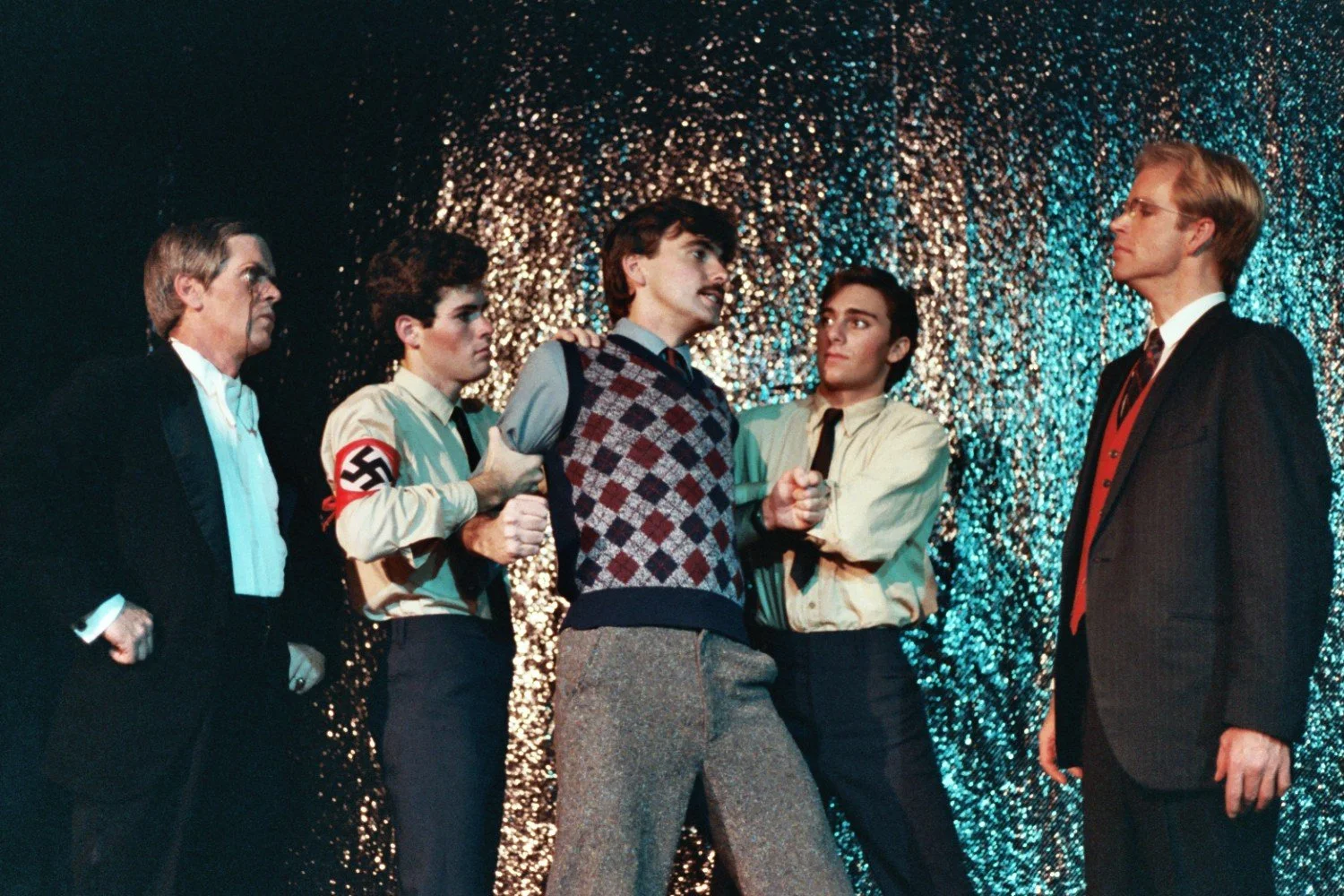 A scene from a theater play with four men, two actors wearing Nazi uniforms, holding a man in a sweater, on a shiny backdrop.