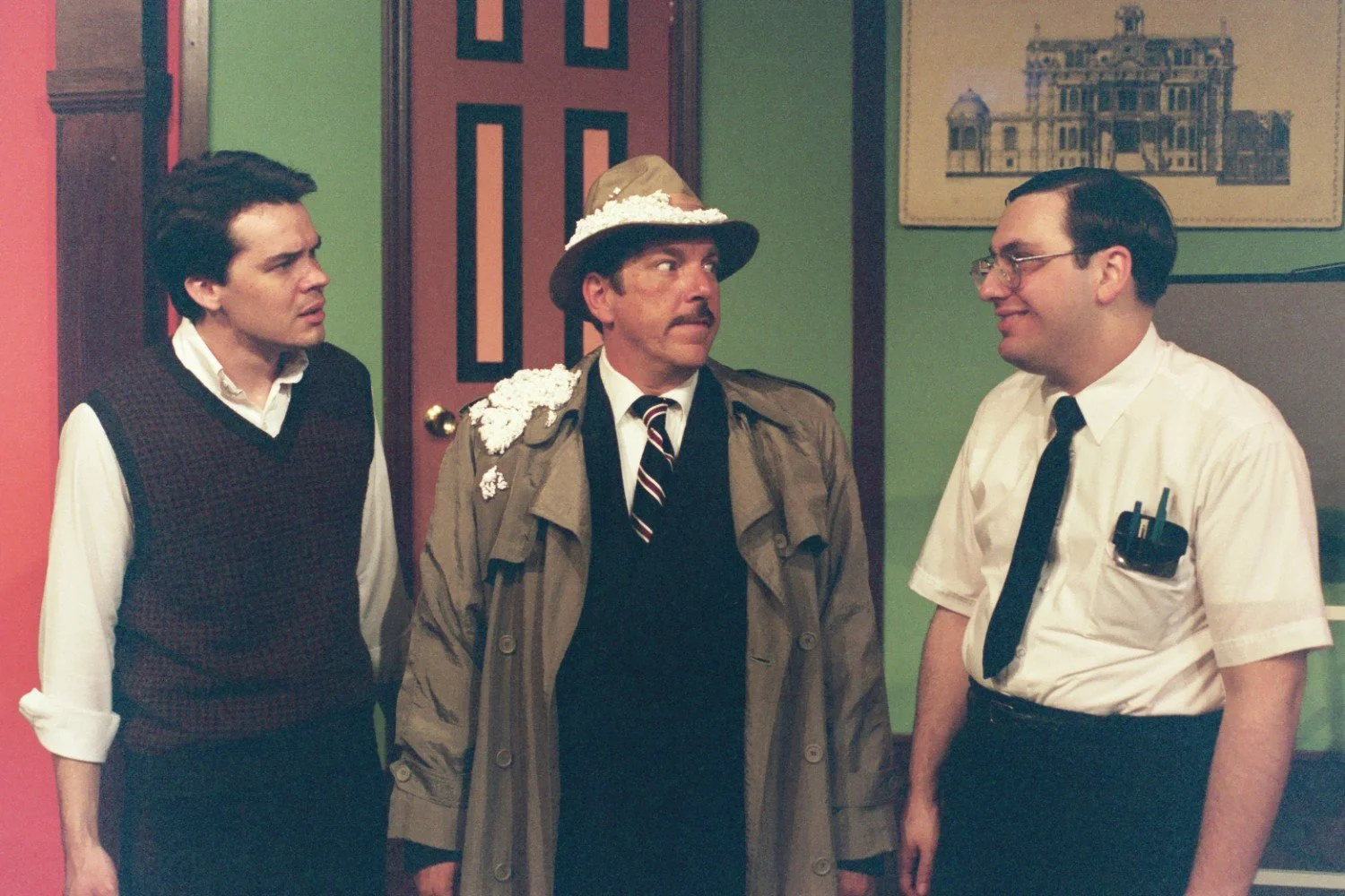 Three men indoors, one with a trench coat and hat covered in shaving cream, standing in front of a green wall.