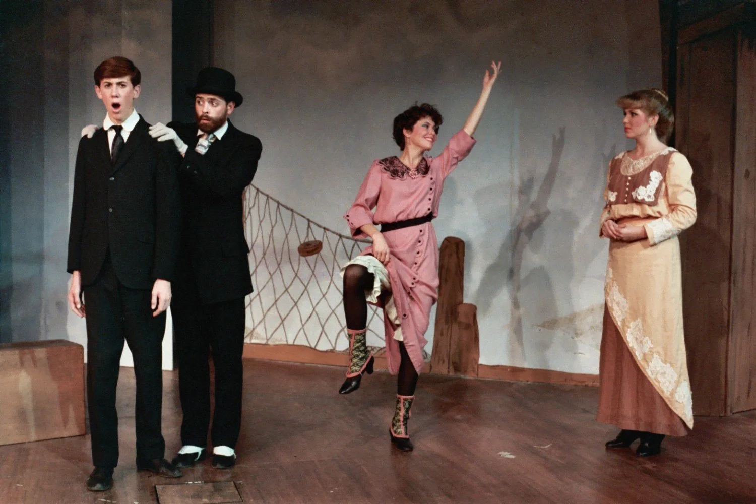 Stage performance with actors in period costumes; one man in a bowler hat adjusts the posture of another man in a suit, while a woman in a pink dress poses with a raised arm and leg, and another woman in a long beige dress watches.