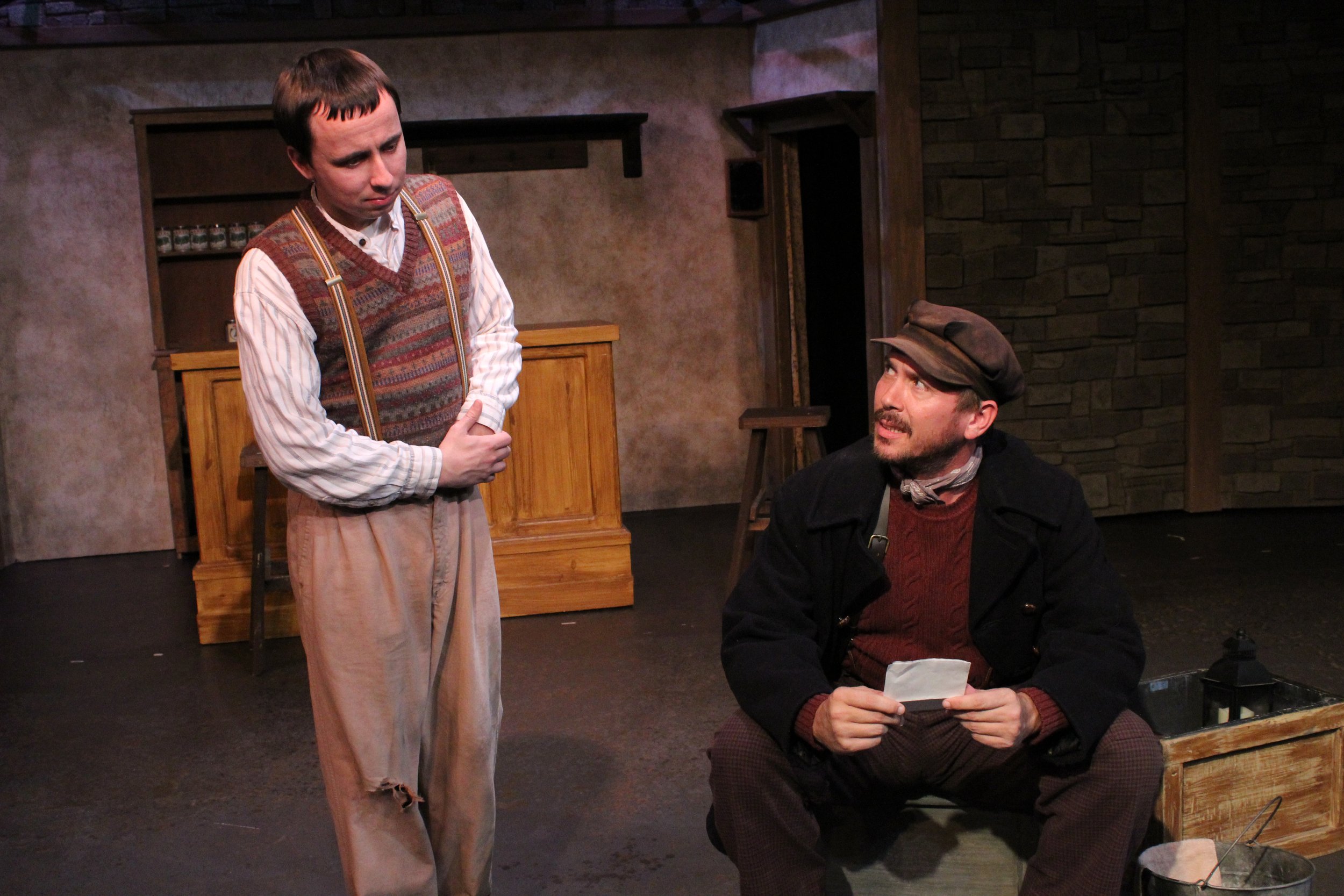Two actors on stage in a theatrical play, one standing wearing a striped shirt and patterned vest, the other sitting wearing a cap and coat, holding a piece of paper, with a rustic set that includes a stone wall and wooden furniture.