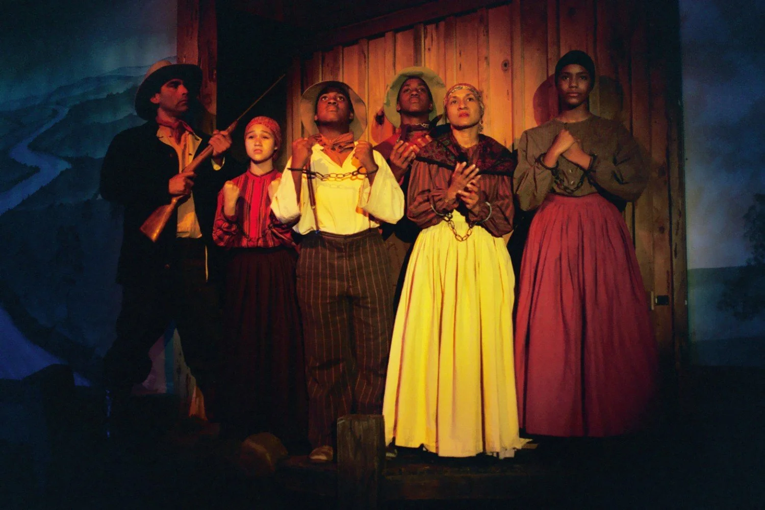 Group of individuals in period clothing with some wearing chains, appearing to portray a historical scene, set against a wooden backdrop.