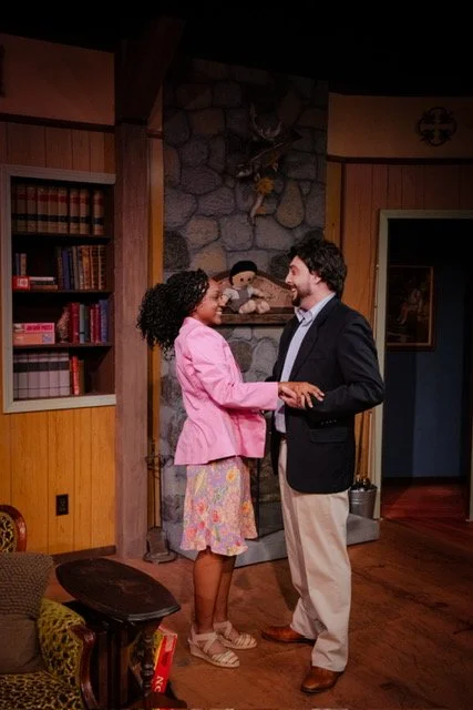 A couple holds hands in a cozy living room with a stone fireplace. The woman is wearing a pink blazer and floral skirt, while the man is in a dark blazer and beige pants. Wooden bookshelves and a decorative wall piece are visible in the background.