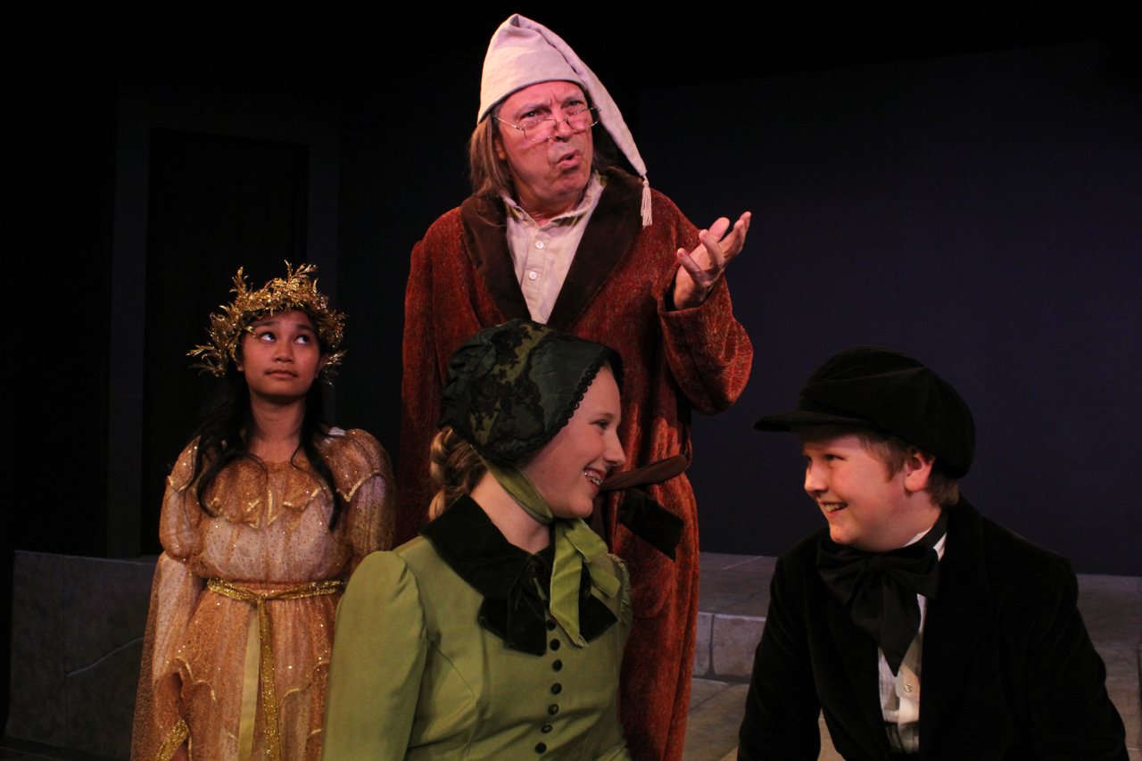 Actors on stage performing a scene from 'A Christmas Carol,' featuring an older man in a nightcap and robe, a young girl in a gold dress with a wreath, and two children in period costumes, smiling.