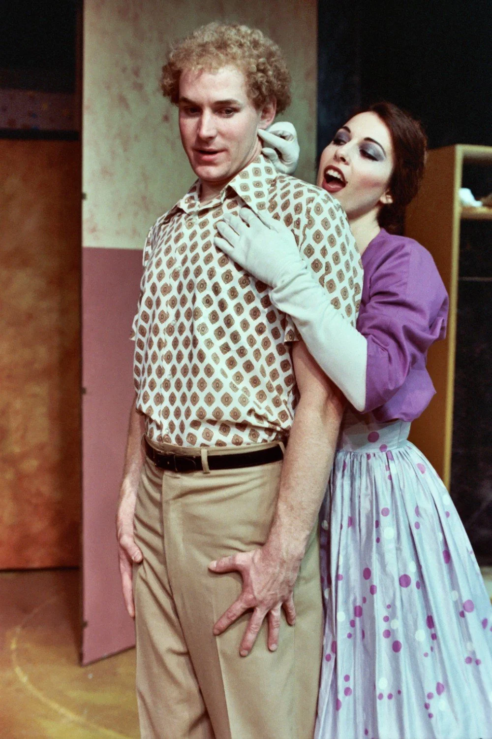 A woman in a purple dress and white gloves embraces a man wearing a patterned shirt and beige pants, in a theatrical setting.