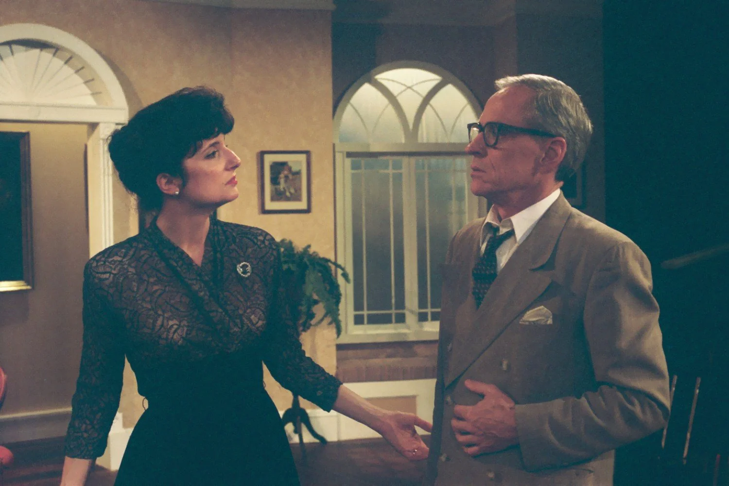 A woman in a black lace dress and a man in a beige suit are having a conversation in a vintage-style room with arched windows and framed art on the walls.