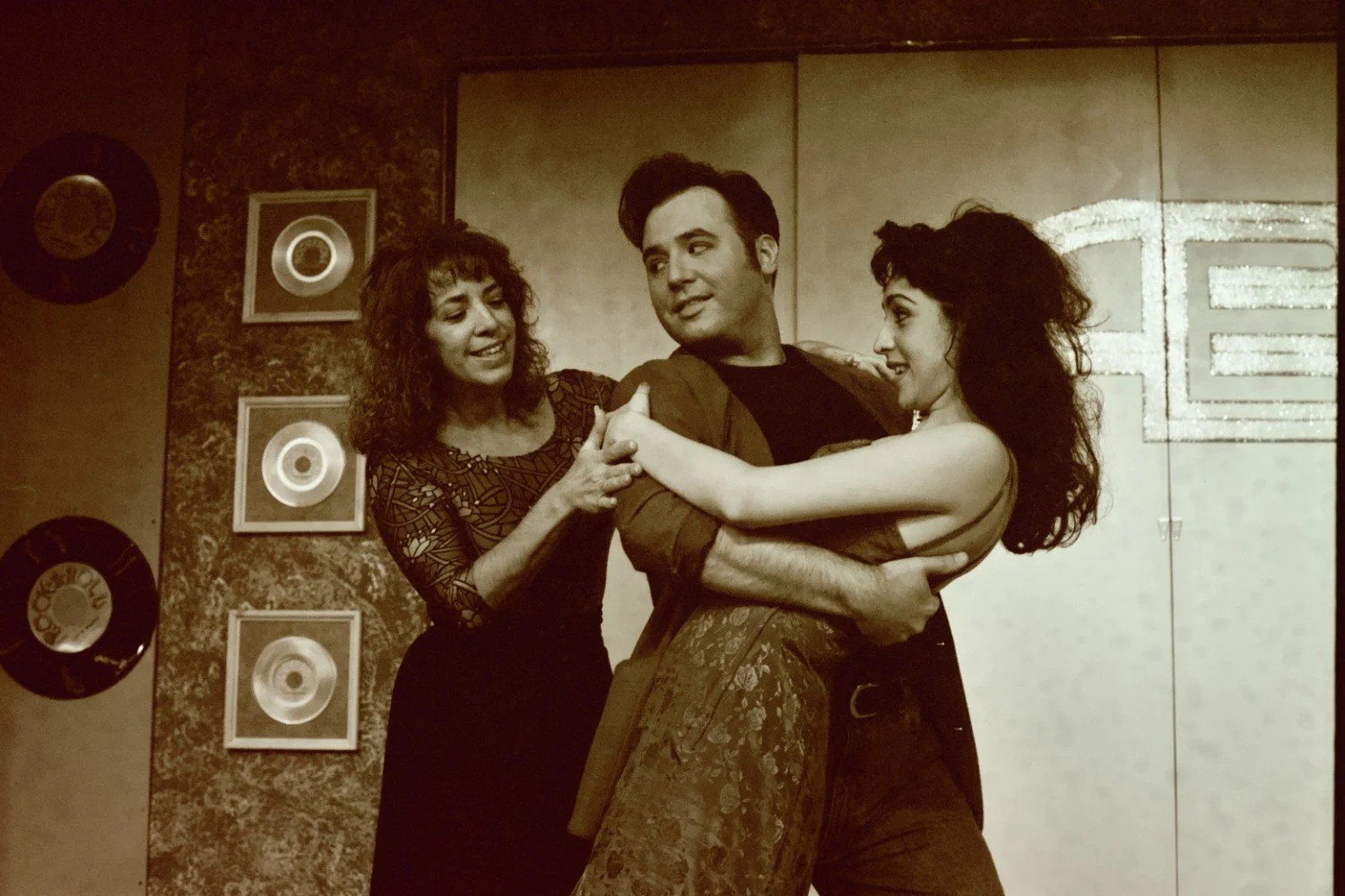 Three people on stage, one man and two women, in a dancing pose with vintage-style decor and records on the wall.