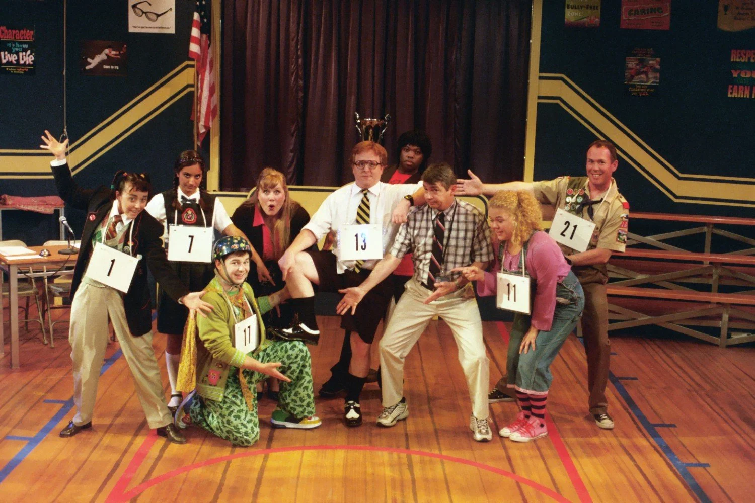 Cast members of a stage production dressed in various costumes, posing energetically on a wooden floor with numbered signs around their necks, suggesting a spelling bee theme. An American flag is in the background.