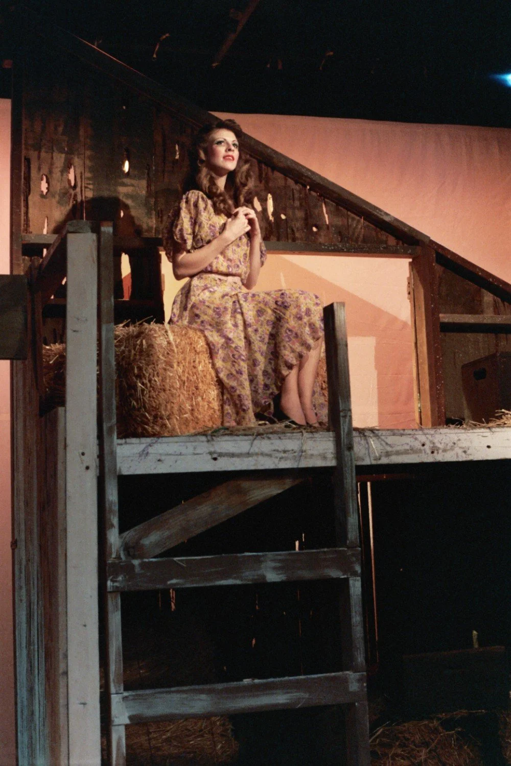Woman in a purple floral dress sitting on hay in a rustic barn setting, looking thoughtful.