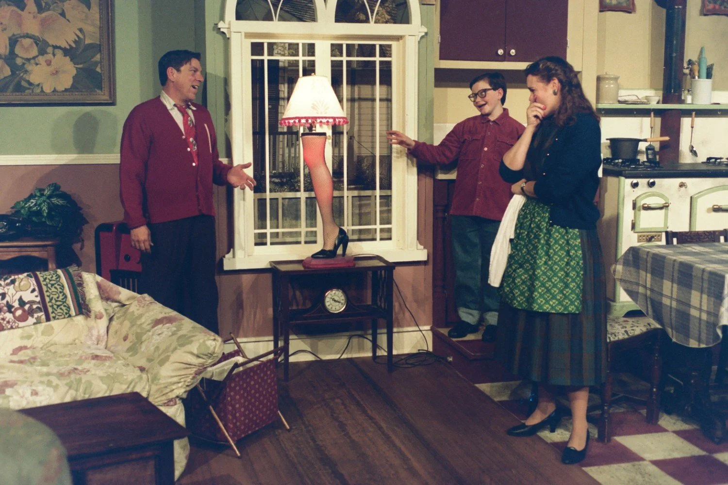 Three people in a living room, one man pointing at a leg lamp in the window, another laughing, and a woman covering her mouth in amusement. The scene appears to be from a vintage-style setting with floral decor. The leg lamp is a prominent feature on
