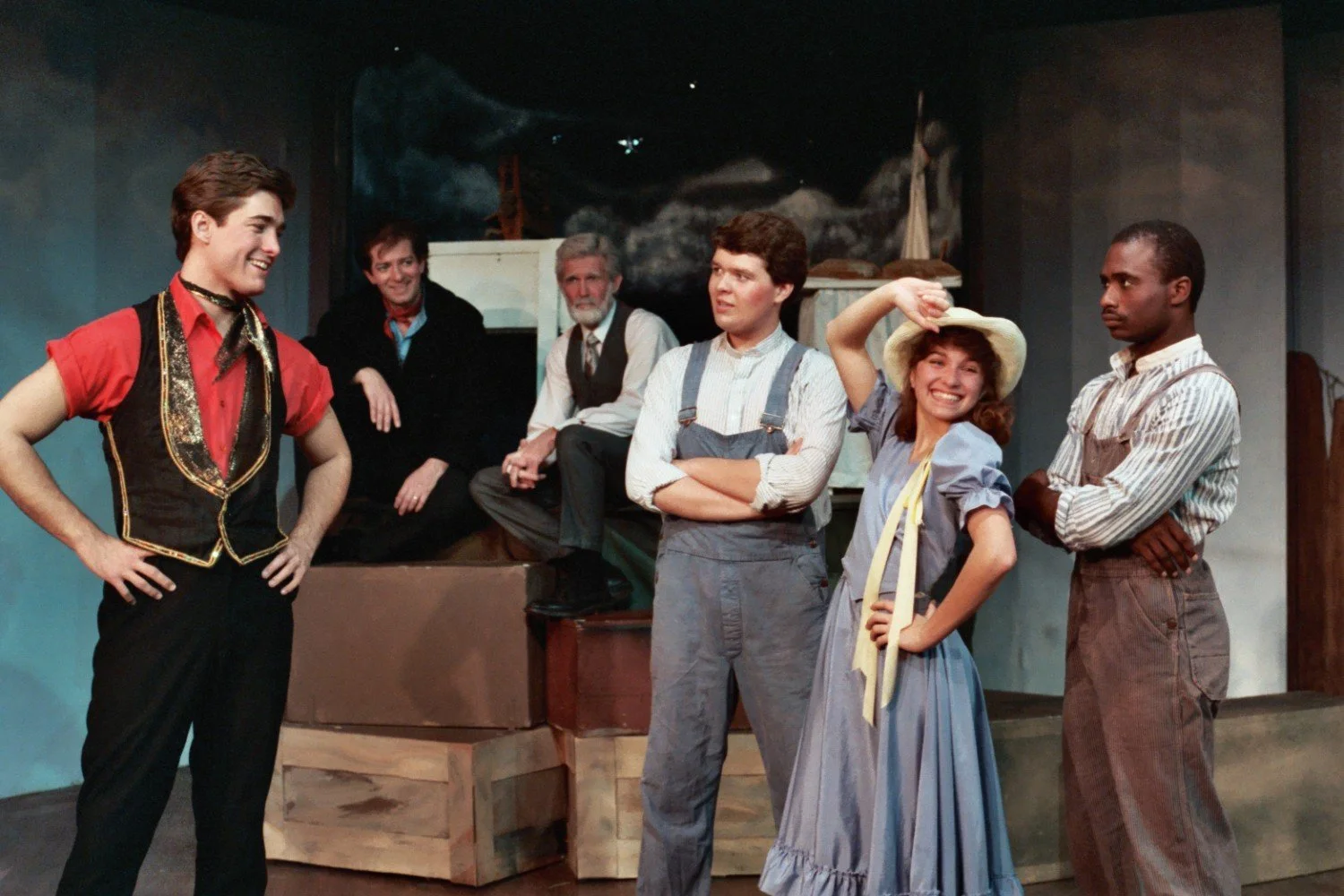 Group of actors on stage in period costumes, including a man in a red shirt and vest, another in overalls, a woman in a bonnet and dress, and two others seated in the background.