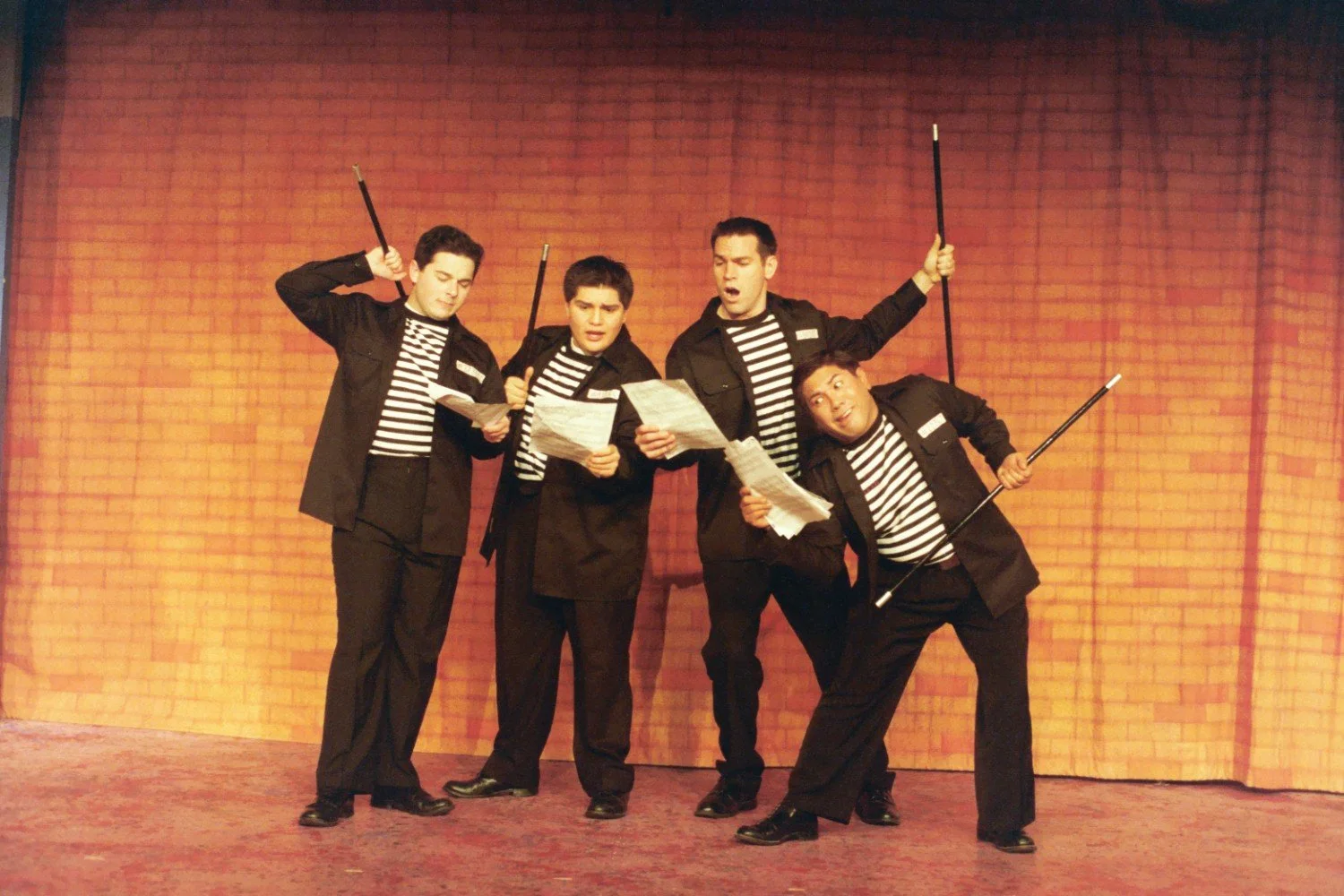 Four men performing on stage, dressed in striped shirts and black jackets, holding papers and canes, standing in front of a brick-patterned curtain.
