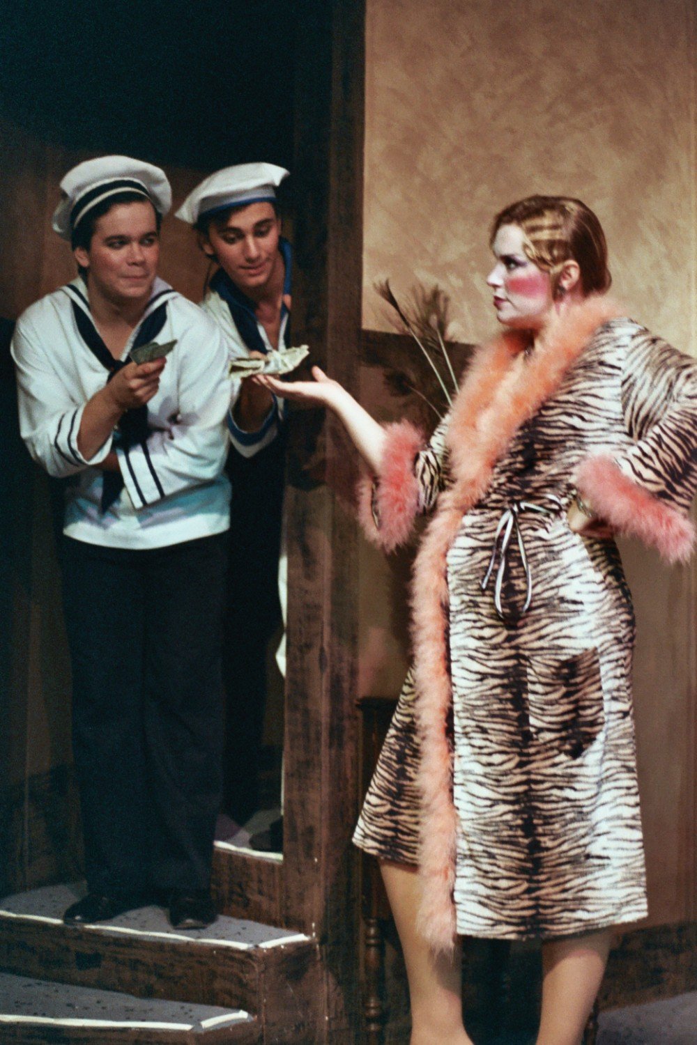 Two men in sailor costumes offering money to a woman in a fur-lined robe on stage.