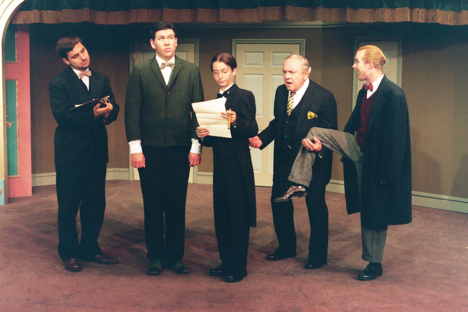 A group of actors on stage in vintage attire performing a comedic scene. The set includes doors and a draped archway, with one actor holding a paper and another with a raised leg.