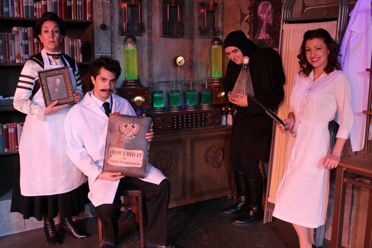 Four people in a vintage laboratory setting with scientific equipment, wearing period costumes. One person holds a book titled "How I Did It" by Victor Frankenstein.