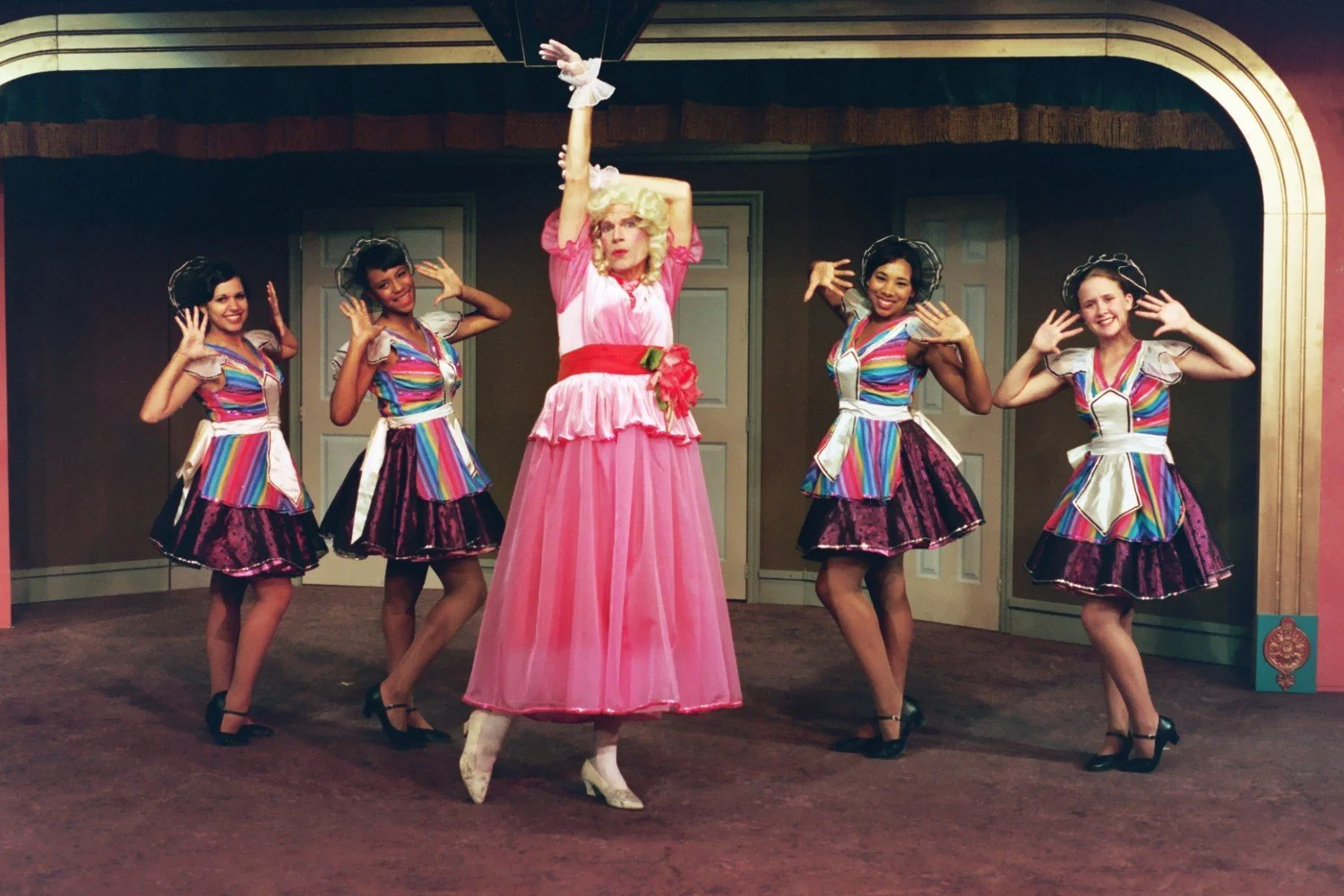 Five people posing on stage in colorful costumes, with one person in a pink dress and the others in rainbow-striped tops and skirts, performing a dance routine.