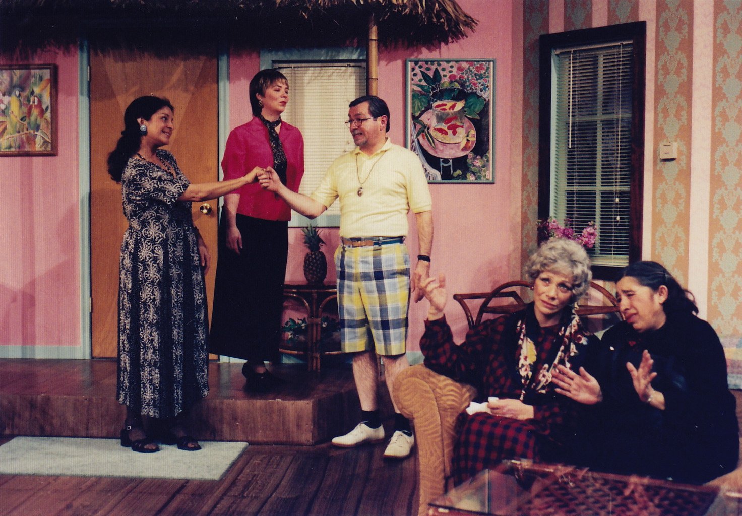 Five people in a living room, some standing, some sitting, engaged in conversation. The room has pink walls, a door, and colorful artwork. One person is shaking another's hand.