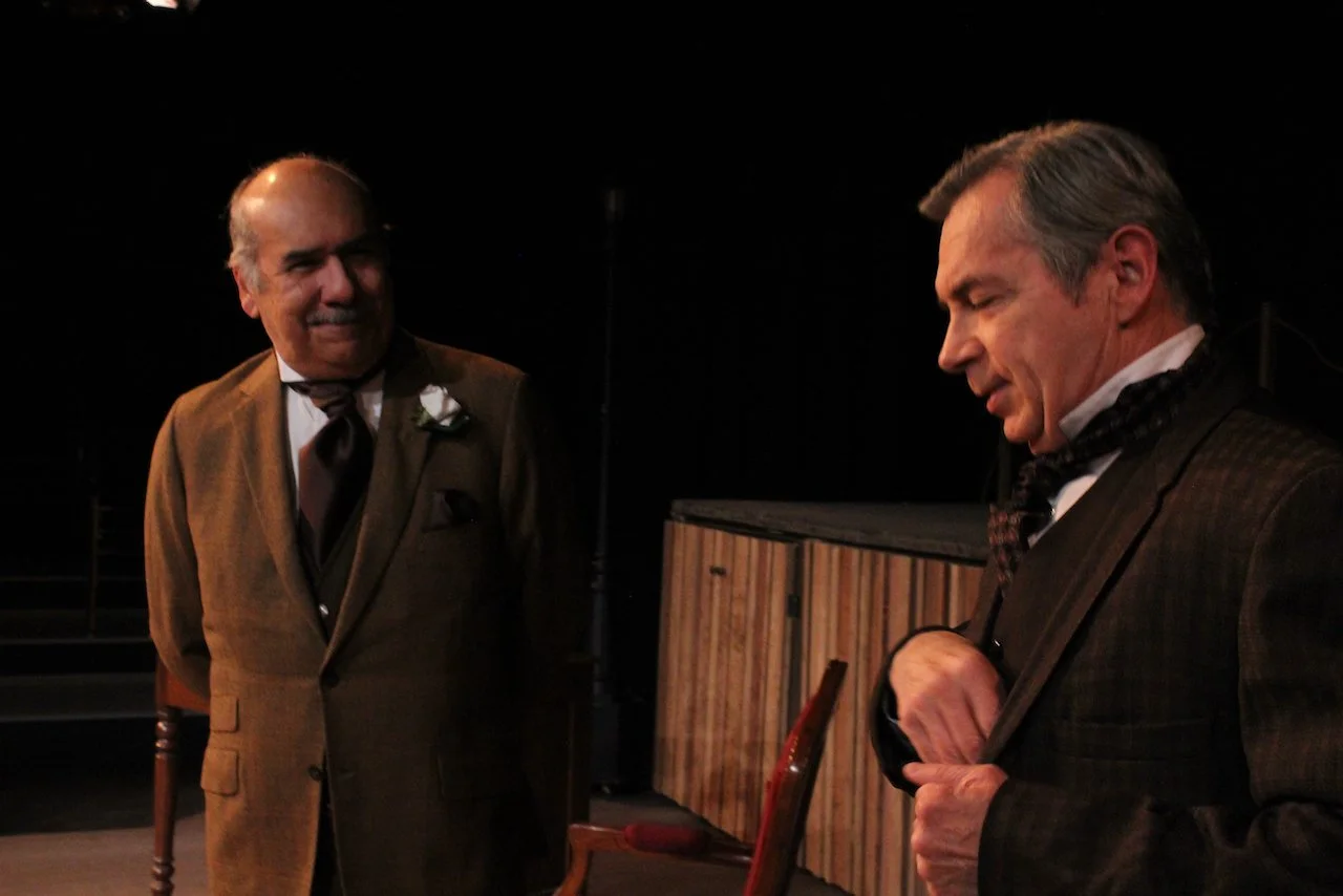Two men in vintage suits on stage, one with a flower in lapel