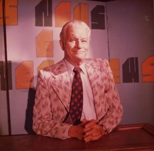 Man in patterned suit standing in front of a colorful "News" sign on a TV set, smiling.
