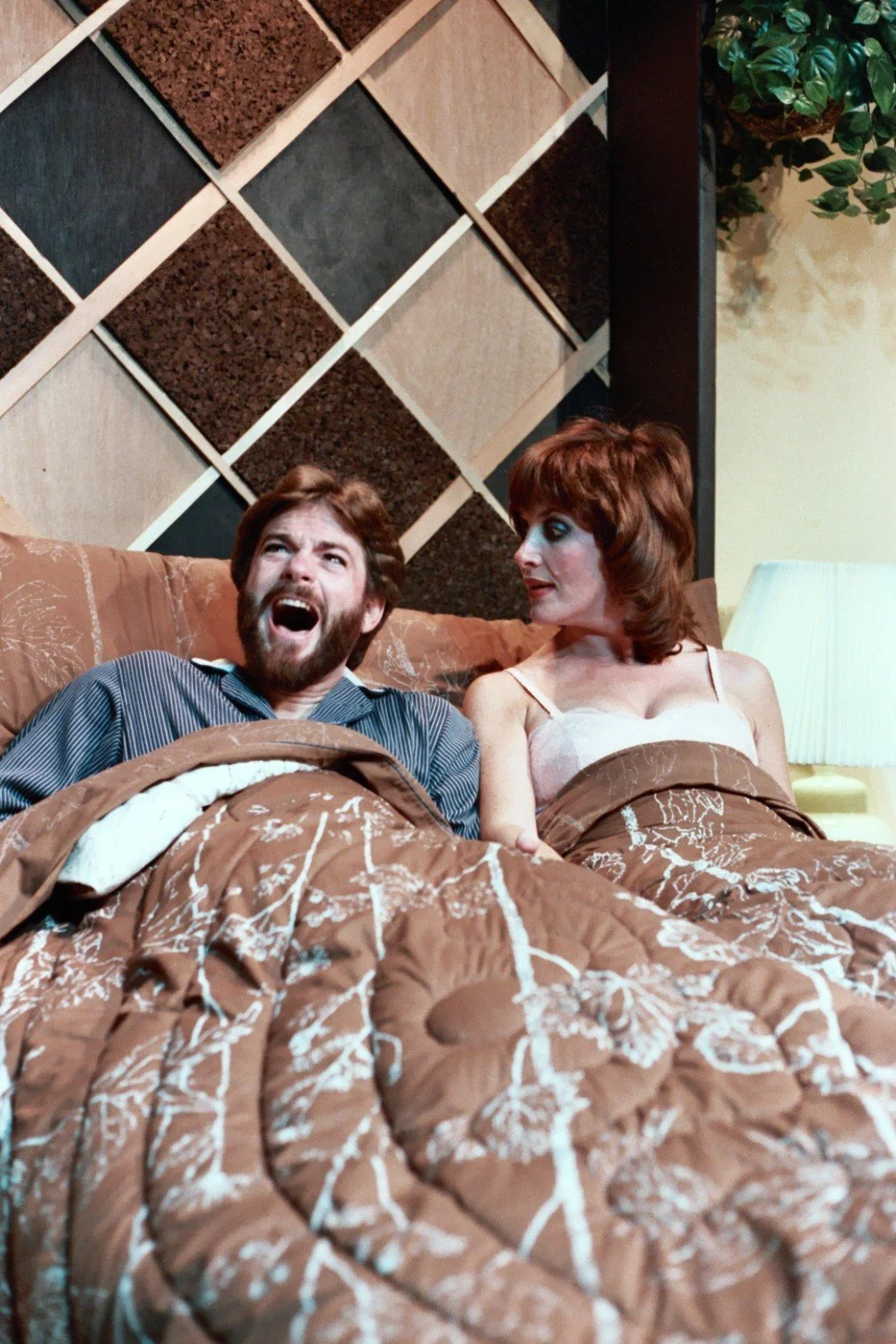 A man and woman in bed, covered by a patterned brown blanket, with a geometric wall design and a lamp in the background. The man appears surprised or shouting, while the woman looks at him.