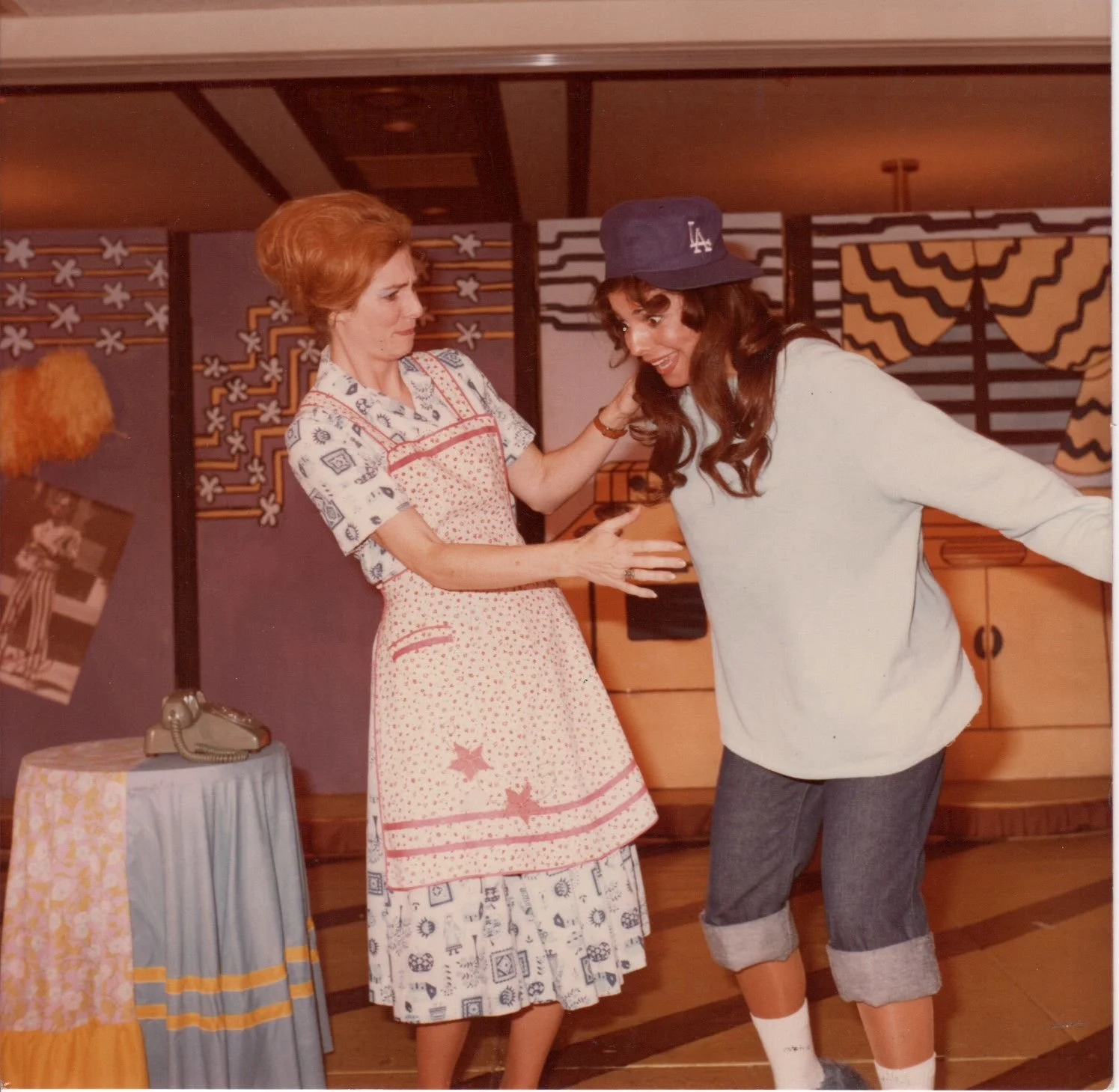 Two women indoors in vintage clothing. One wears an apron and dress, while the other has a casual outfit with a Los Angeles Dodgers cap. They're interacting playfully. Background includes patterned walls, a table with a telephone, and retro decor.