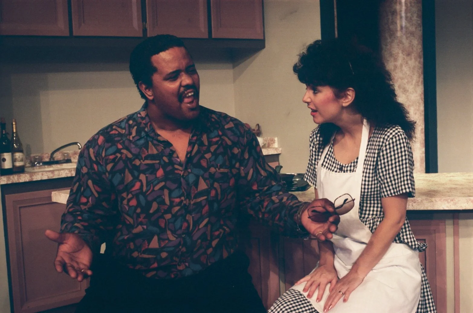 Two people in a kitchen setting, one holding glasses, engaged in conversation.