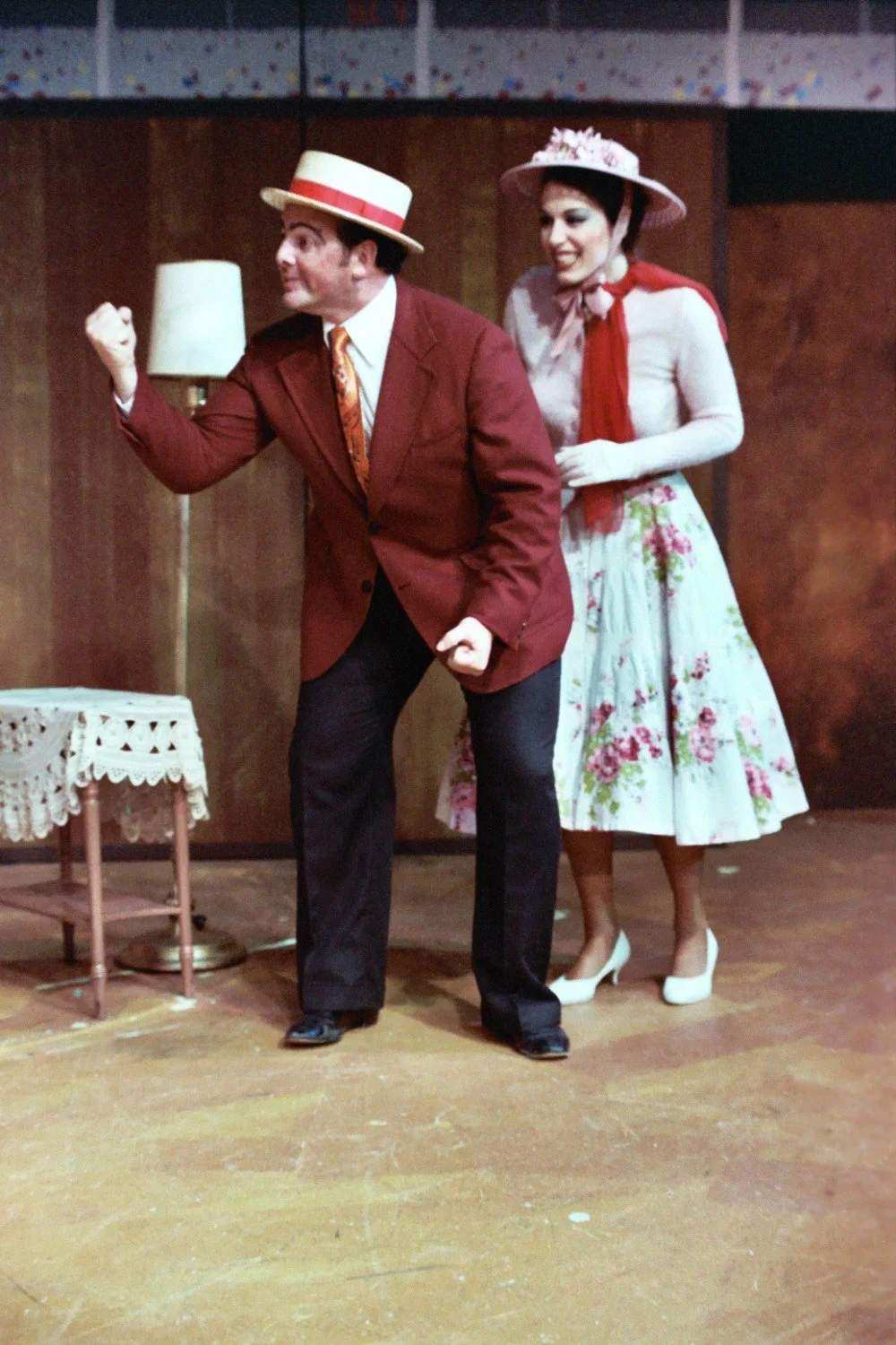 Two people in vintage theater costumes performing a scene on stage. One person is wearing a maroon jacket and straw hat, gesturing with a fist, while the other wears a floral skirt and hat, smiling.
