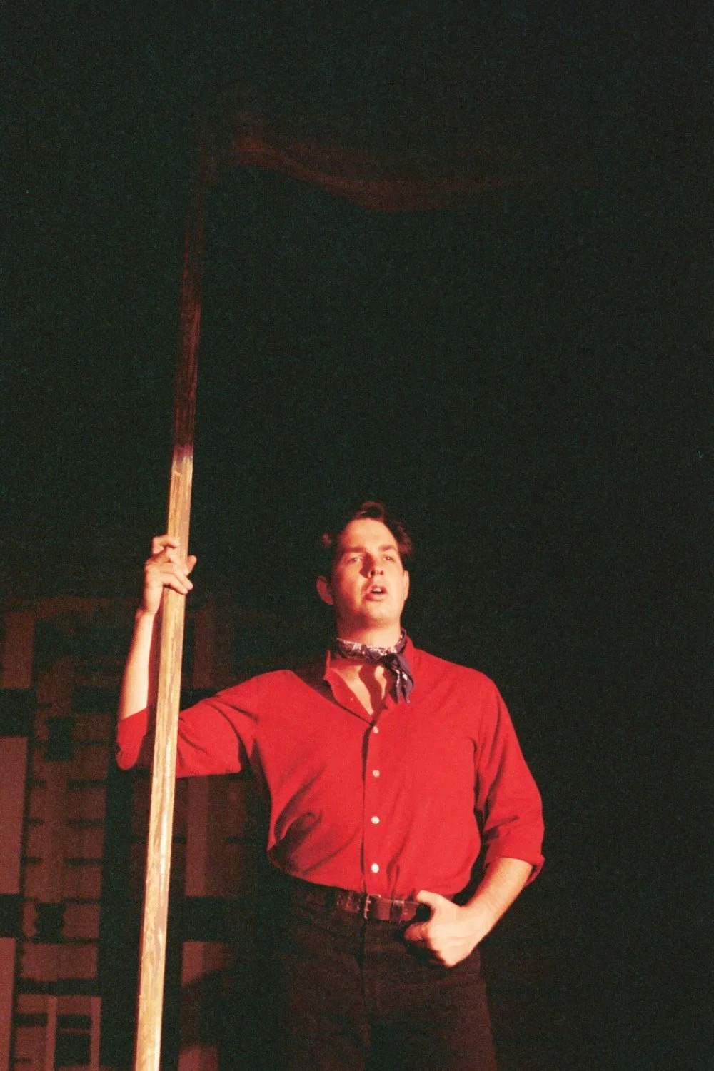 Person in red shirt holding a scythe, performing on stage.
