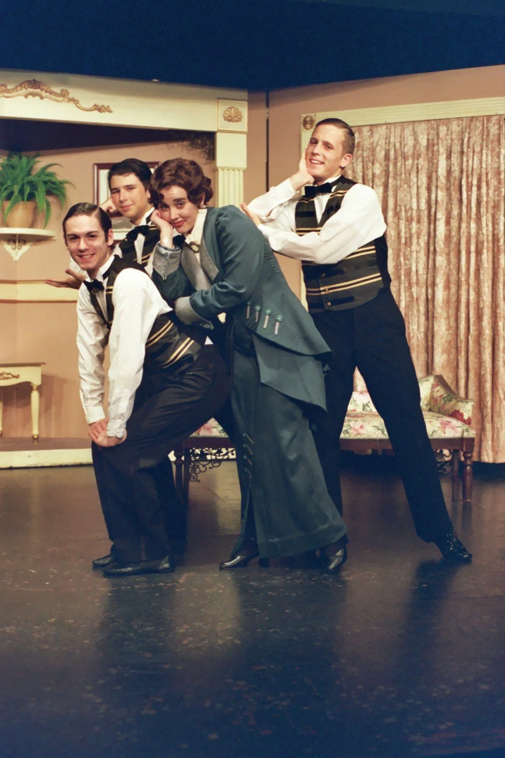 Four people posing on stage during a theatrical performance, with one wearing a long green dress and the others in black and white costumes with vests. The background features a set designed as a parlor with a plant and curtains.