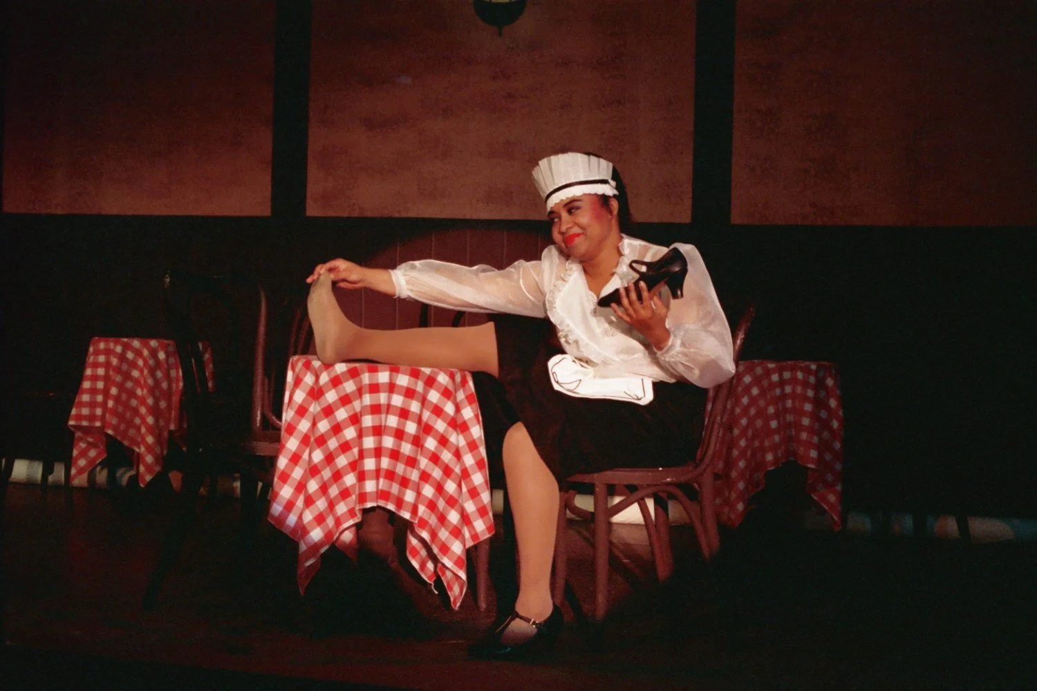 Person wearing a maid's outfit sitting on a chair, stretching their leg on a table with a red and white checkered tablecloth, holding a black shoe.