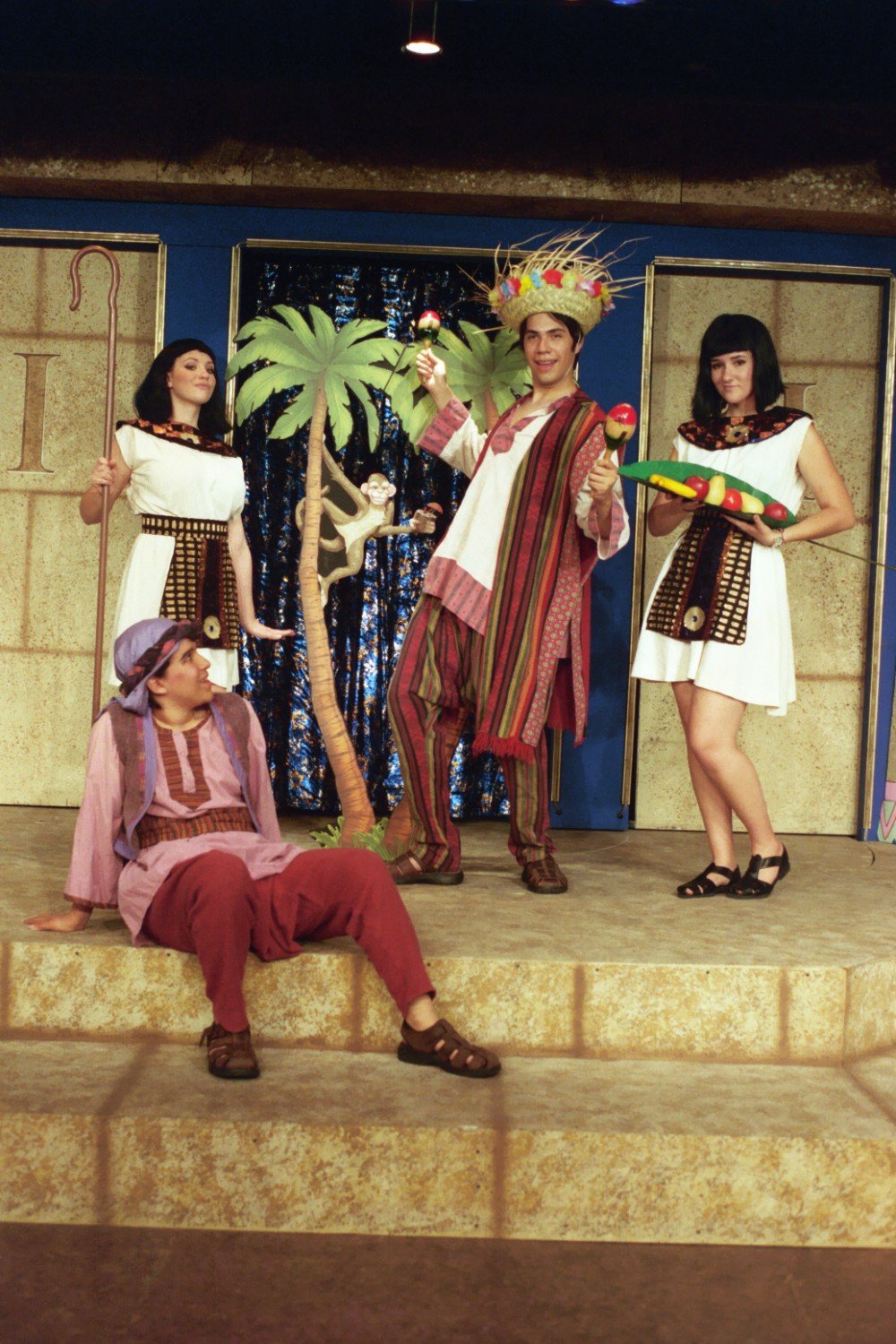 A theatrical scene featuring four actors in colorful costumes on a stage. One actor is seated on the steps wearing a purple headscarf and maroon pants. A standing actor in the center wears a straw hat with maracas, with a fake palm tree beside them. 