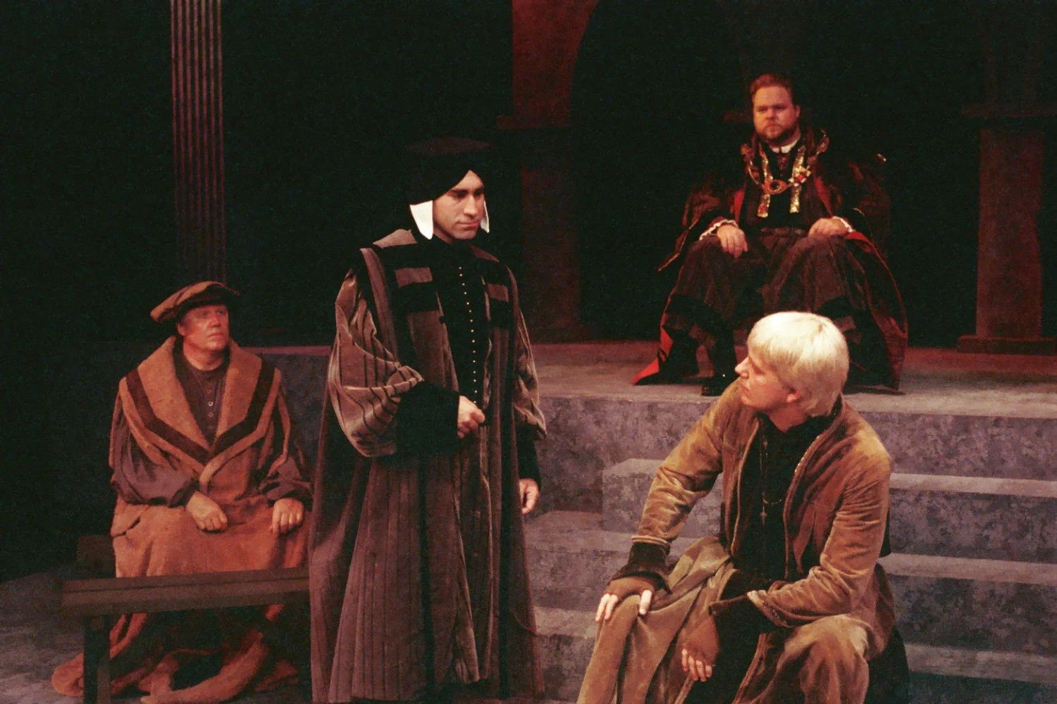 Cast in historical costumes performing a play on stage, with one seated in regal attire, others in robes, in a dramatic scene.