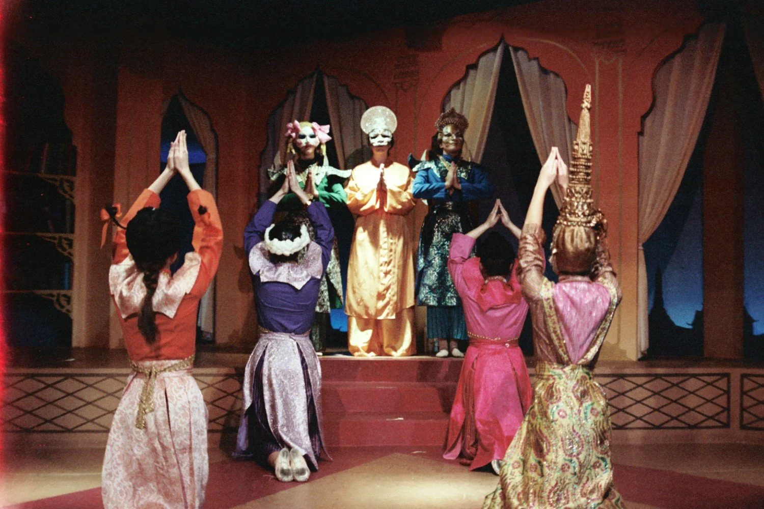 Group of performers in colorful costumes and masks on a stage with raised hands in a cultural or theatrical setting.