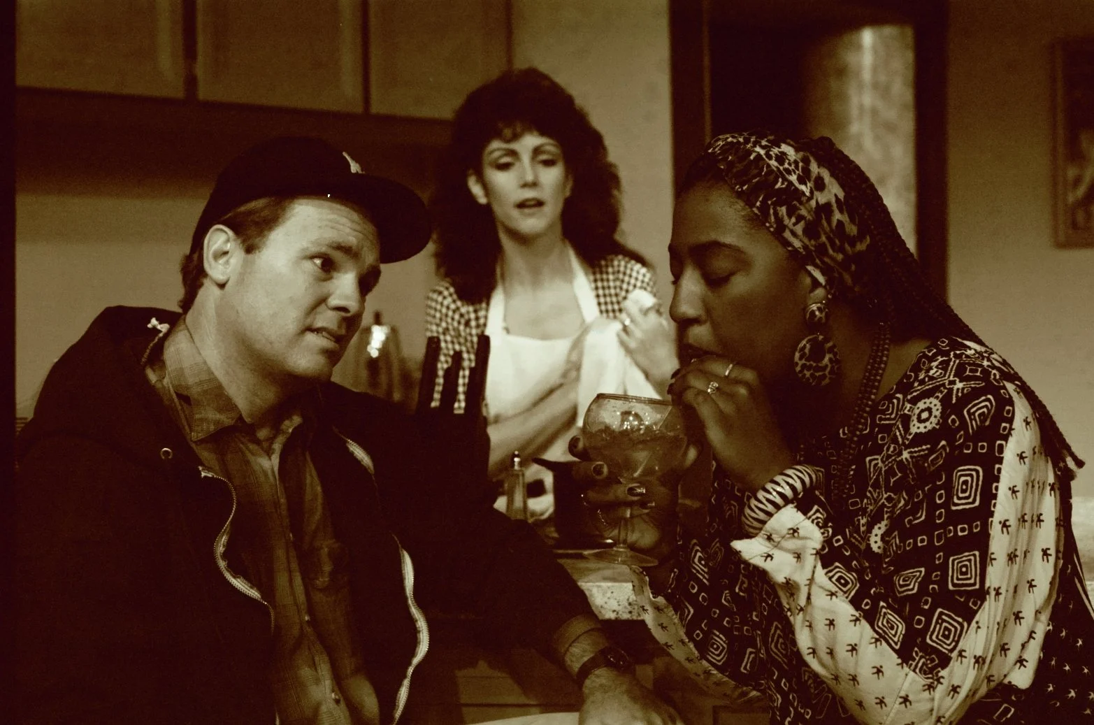 Three people in a dimly lit room; one man wearing a cap is sitting, a woman in an apron is standing behind them, and another woman with braided hair is drinking from a glass. The scene is in sepia tone.