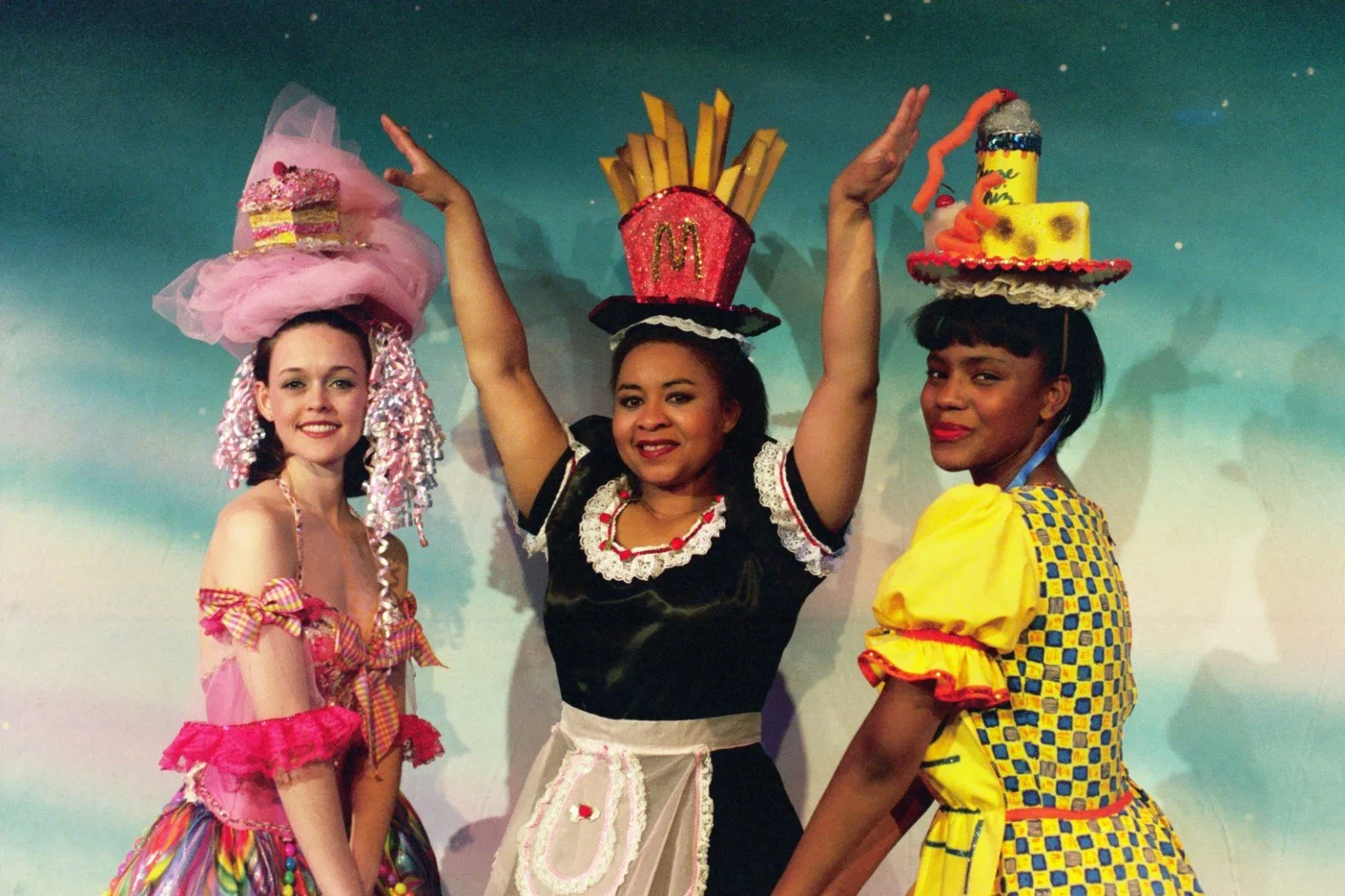 Three women in colorful costumes, one with a cake hat, one with a fries hat, and one with a food-themed hat, posing in front of a blue background.