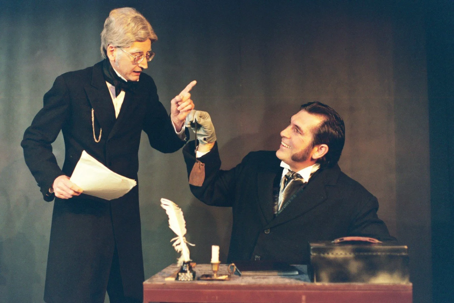 Two men in period clothing performing a theatrical scene; one standing and holding a paper, the other sitting and smiling with a money bag. A desk with a quill and inkwell is in front of them.