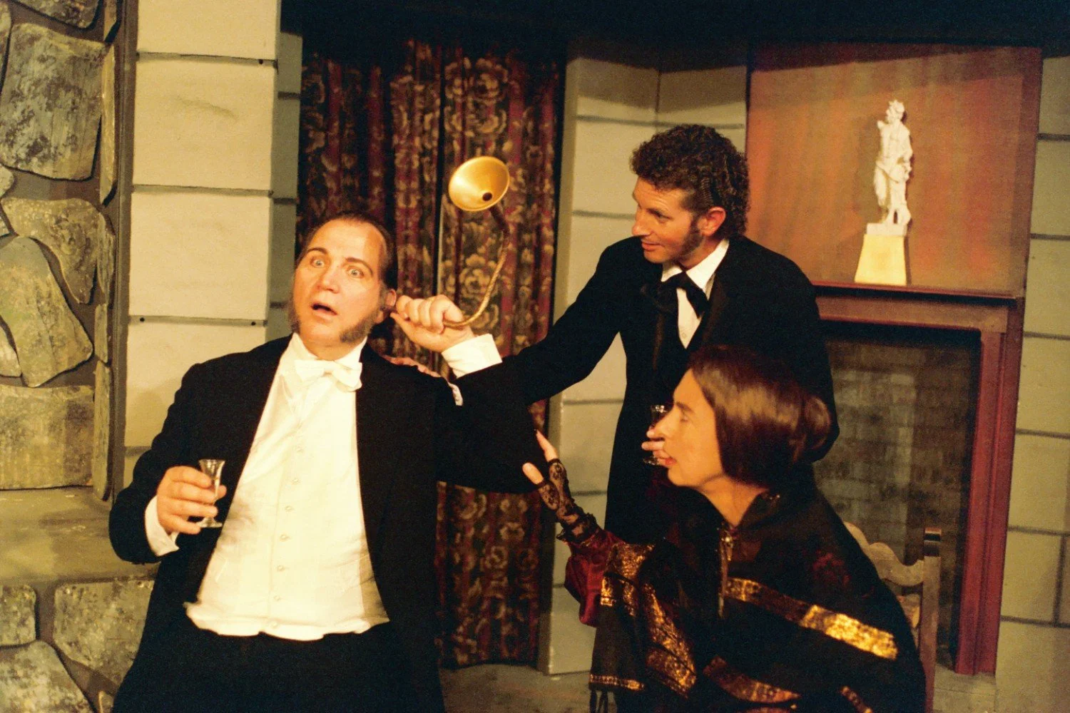 Three people in Victorian-era attire, one holding a hearing trumpet, in a theatrical setting.