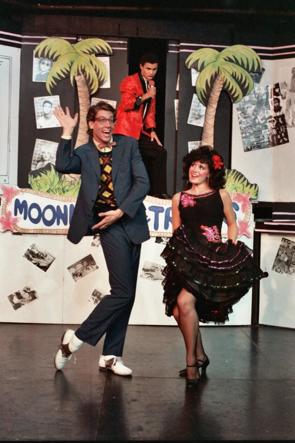 Three performers in a retro-themed stage show; one male in a suit and bow tie posing in front, a female in a black dress with red accents dancing beside him, and another male in a red jacket with a microphone in the background. The backdrop features 
