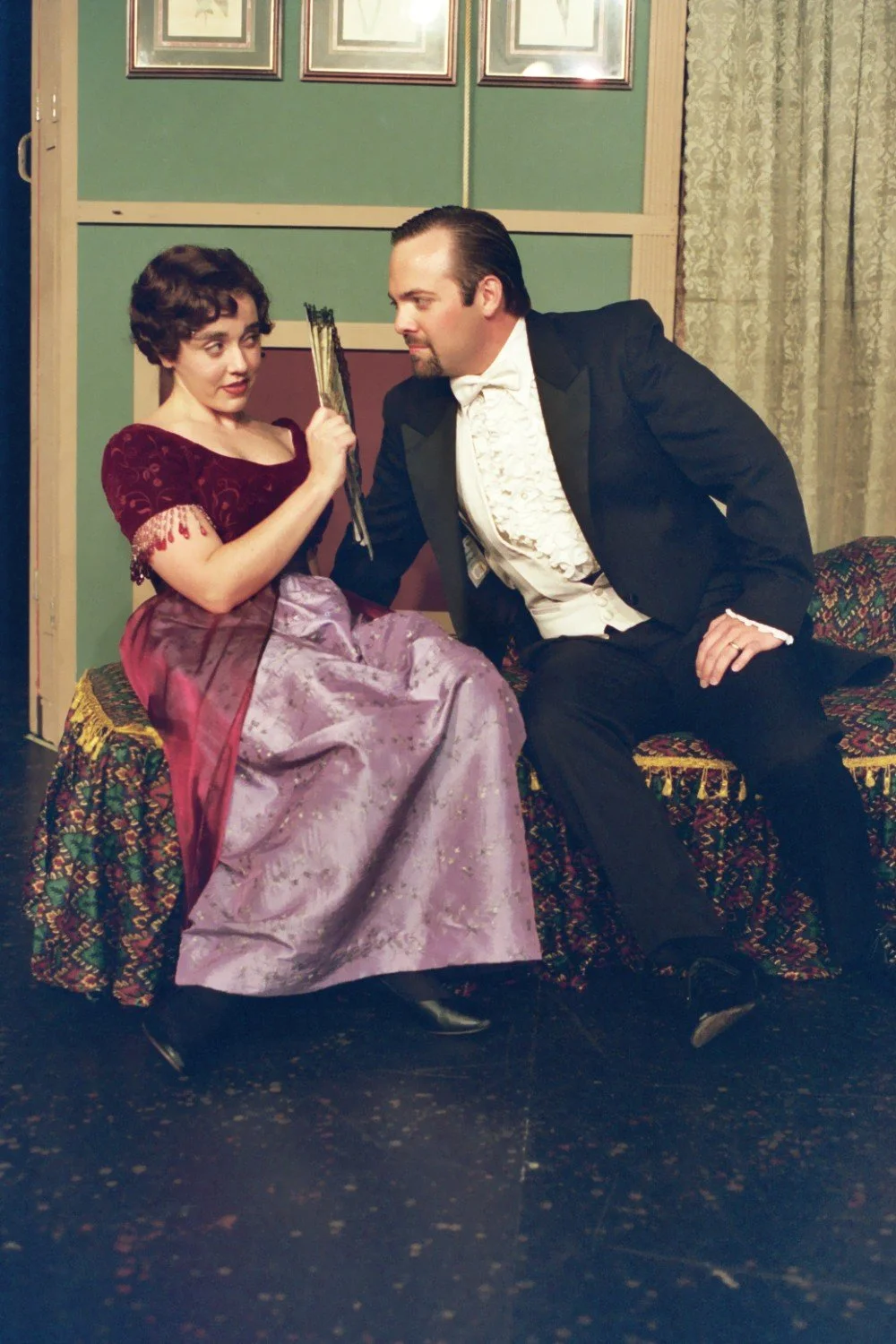 A woman in a Victorian-style gown sitting on a patterned bench, holding a fan, looks at a man in a tuxedo facing her.