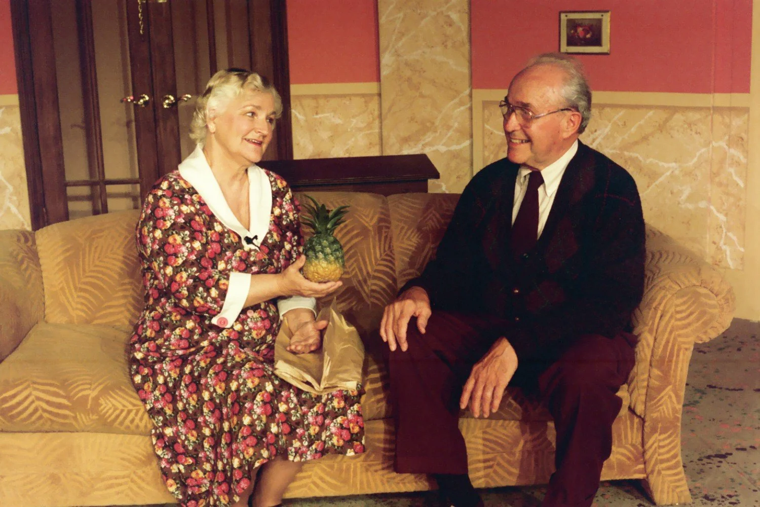 Elderly woman and man sitting on a sofa, woman holding a pineapple.