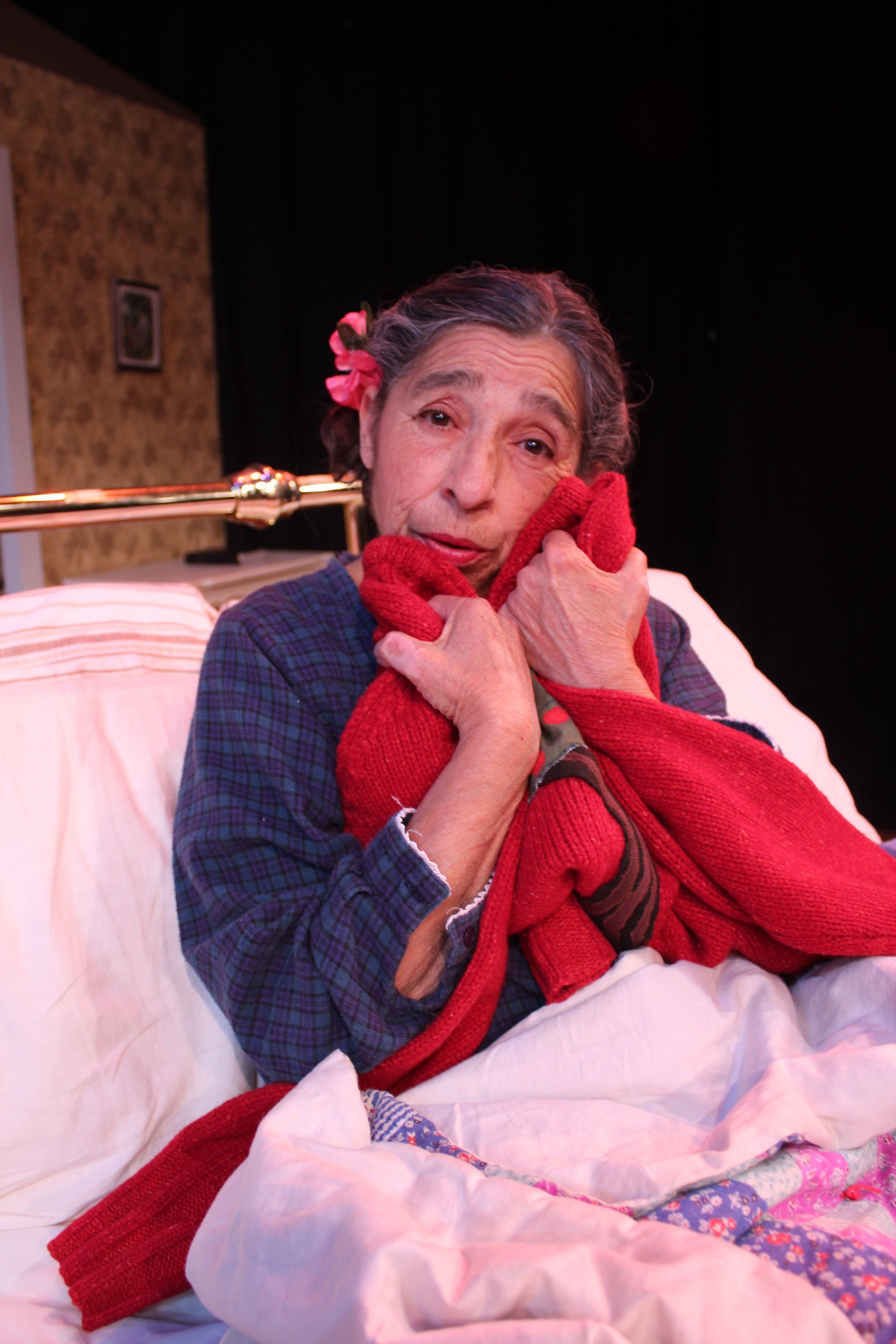 Elderly woman with flower in her hair hugging a red blanket, seated in bed.